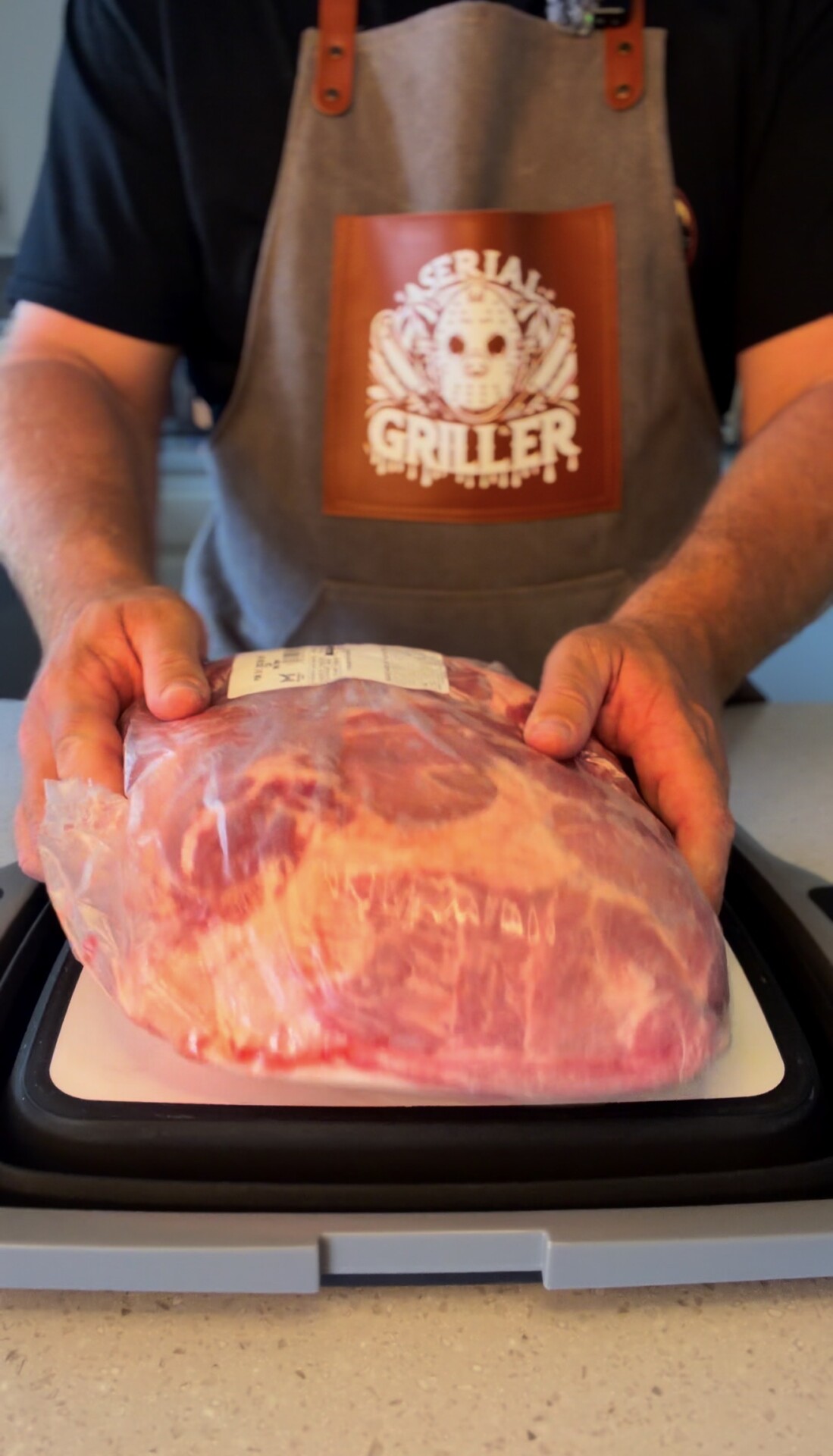 A person wearing an apron labeled Serial Griller holds a large, raw cut of meat wrapped in plastic on a cutting board.