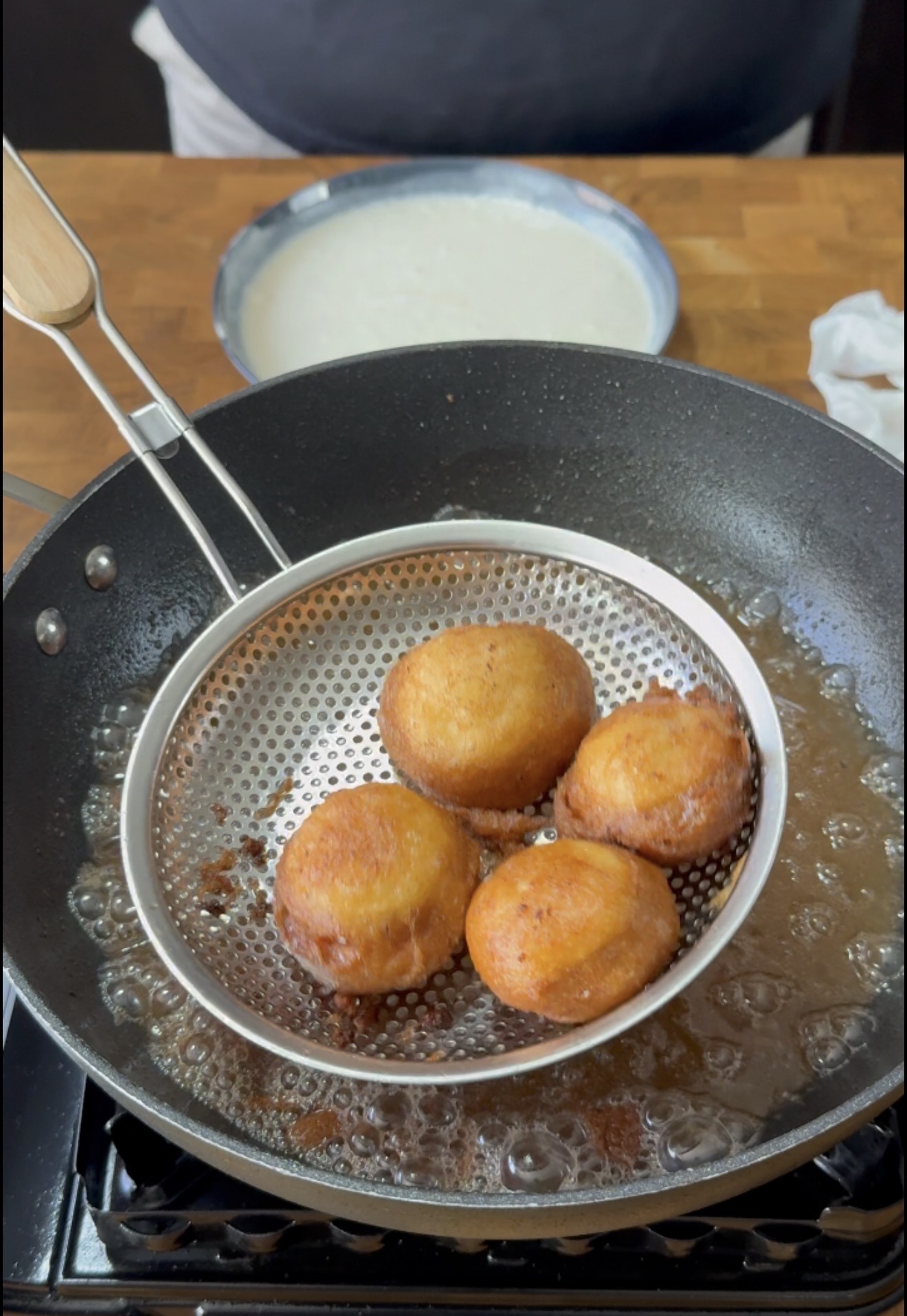 A metal strainer holds three golden brown fried balls over a black pan with hot oil. A plate with white batter and a wooden surface are visible in the background.