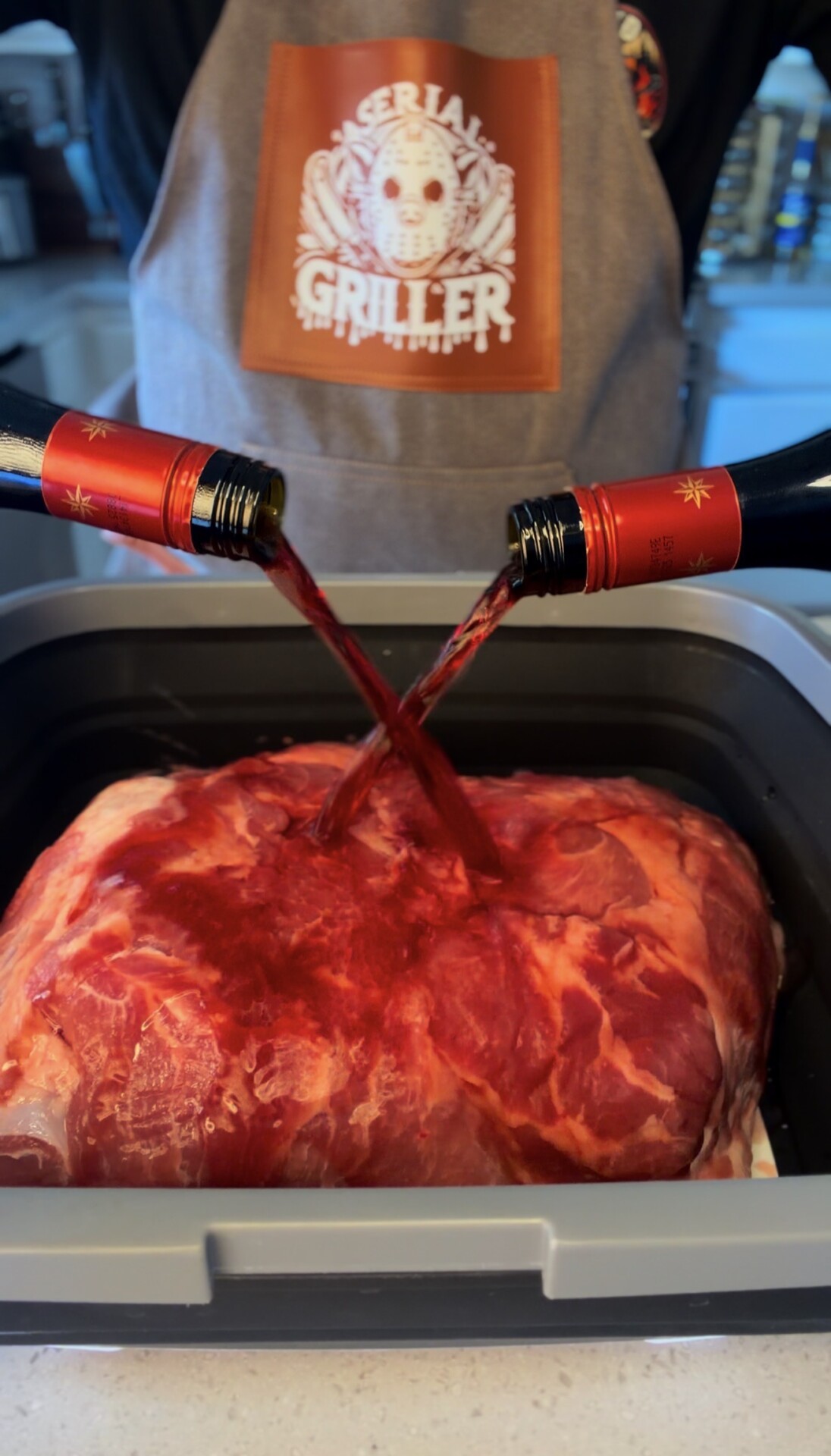 A person in a Serial Griller apron pours red wine from two bottles over a large piece of raw meat in a tray, preparing it for marinating.