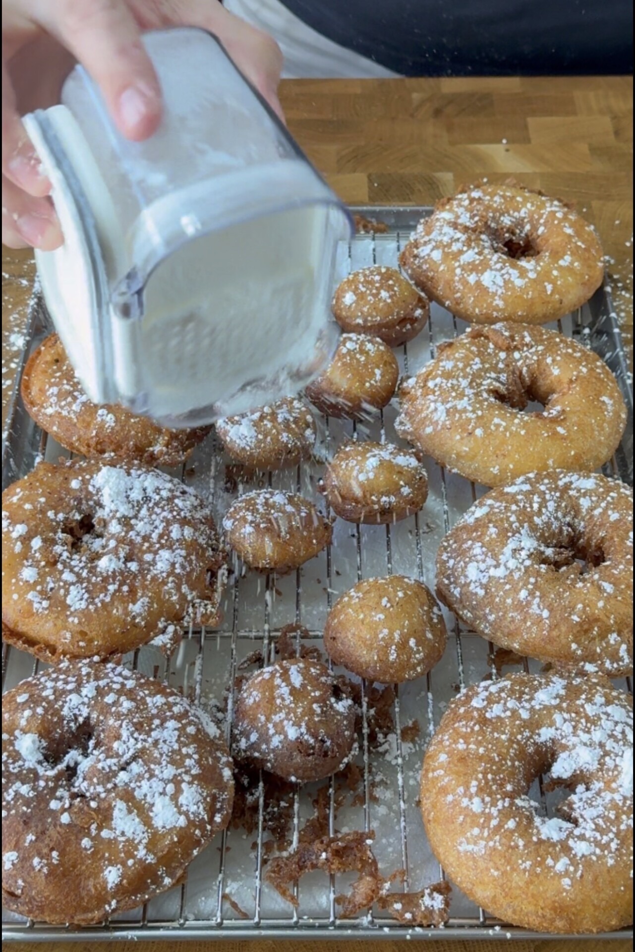 A hand sprinkles powdered sugar from a container onto freshly fried donuts and donut holes, which are cooling on a metal rack atop a wooden surface.