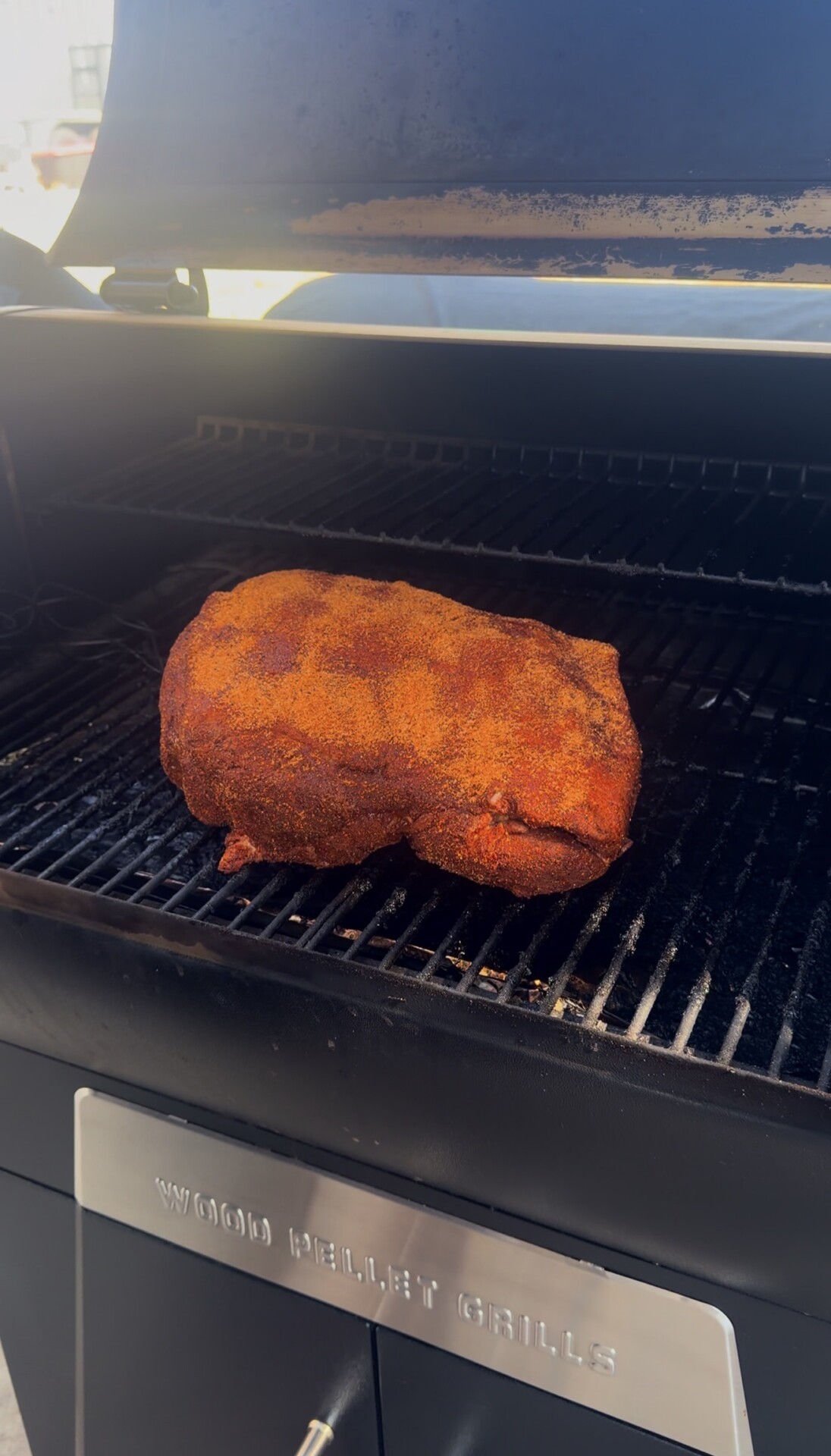 A large, seasoned piece of meat is smoking on the grill grates inside a wood pellet grill, with the grill lid partially open.