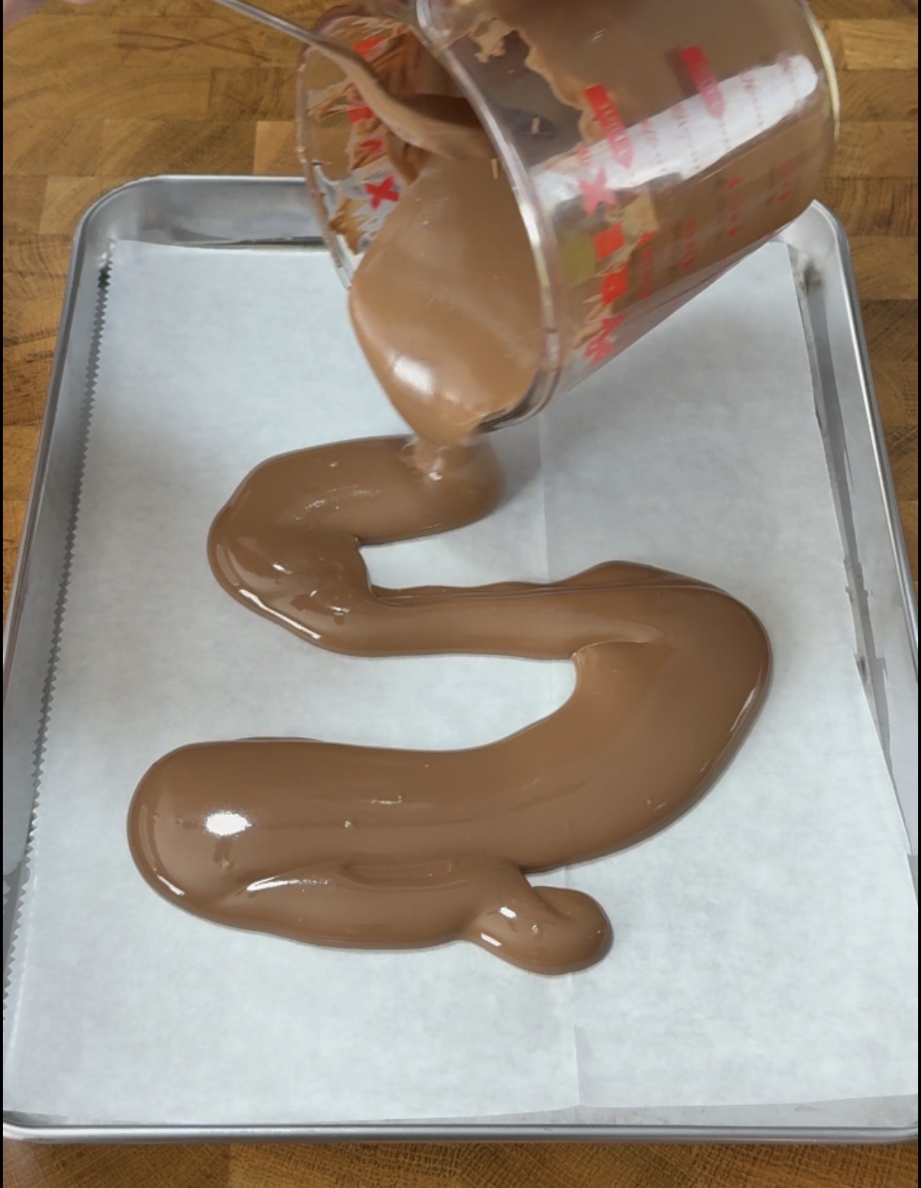 A measuring cup pours melted milk chocolate in a zigzag pattern onto a parchment-lined baking sheet on a wooden surface.