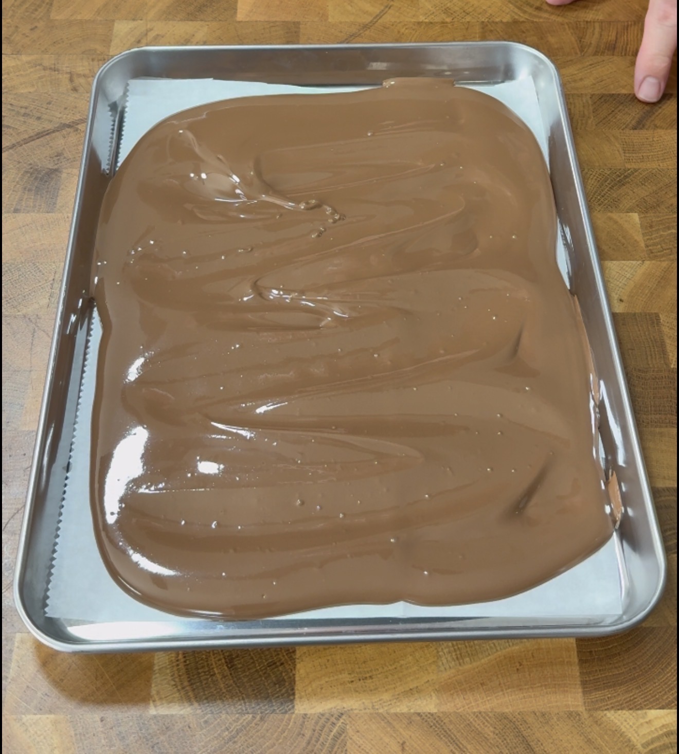 A baking tray on a wooden surface holds a layer of smooth, melted chocolate spread over parchment paper, with a hand visible in the upper right corner.