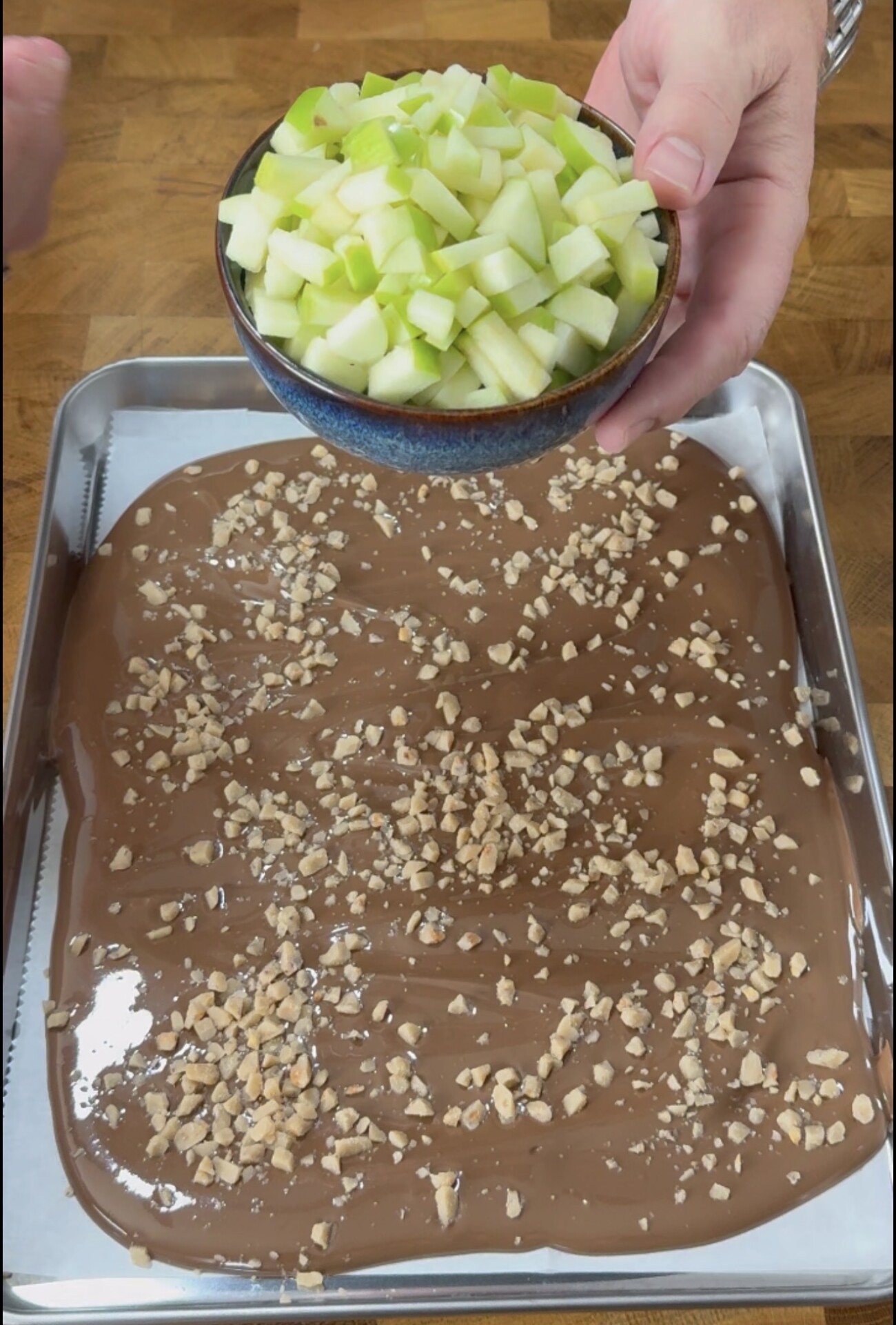 A hand holds a bowl of diced green apples above a baking tray covered with melted chocolate and sprinkled with chopped nuts on a wooden surface.