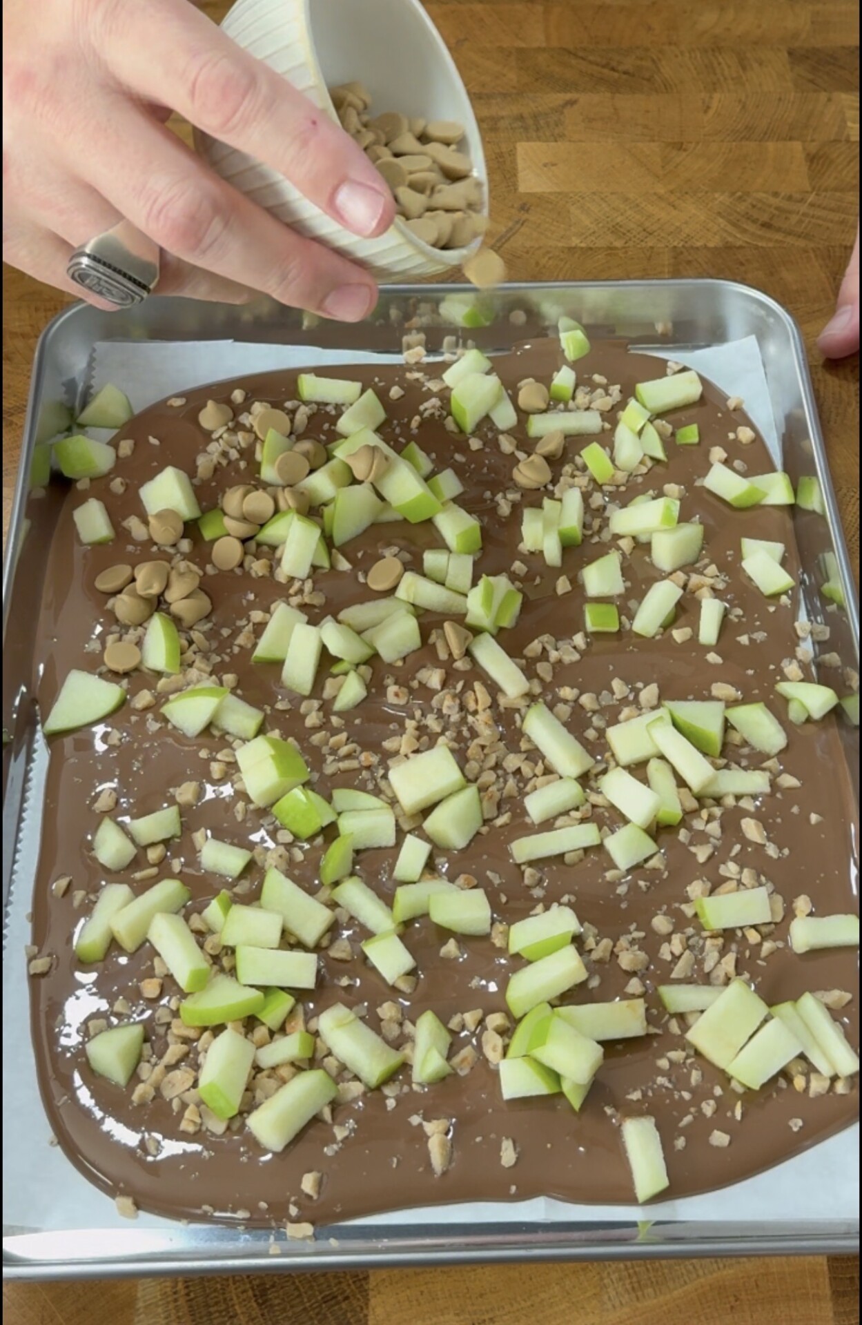 A hand sprinkles nuts onto a baking sheet covered with melted chocolate, chopped green apples, and toffee bits. The sheet is on a wooden surface.