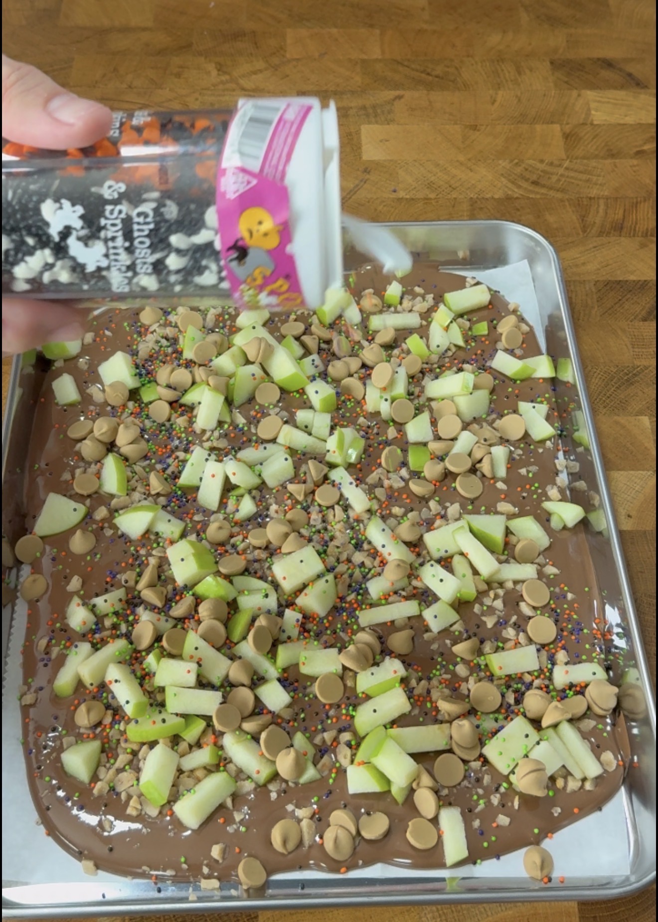 A hand sprinkles Halloween-themed decorations onto a baking sheet covered with melted chocolate, chopped green apples, colorful sprinkles, and caramel chips on a wooden surface.
