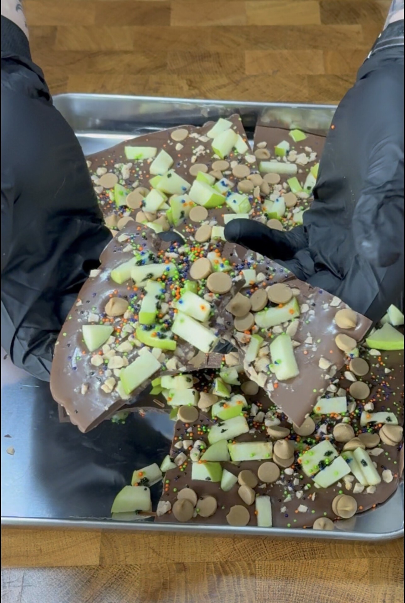 Gloved hands hold pieces of chocolate bark topped with green apple chunks, peanuts, caramel chips, and colorful sprinkles on a metal tray, set on a wooden surface.