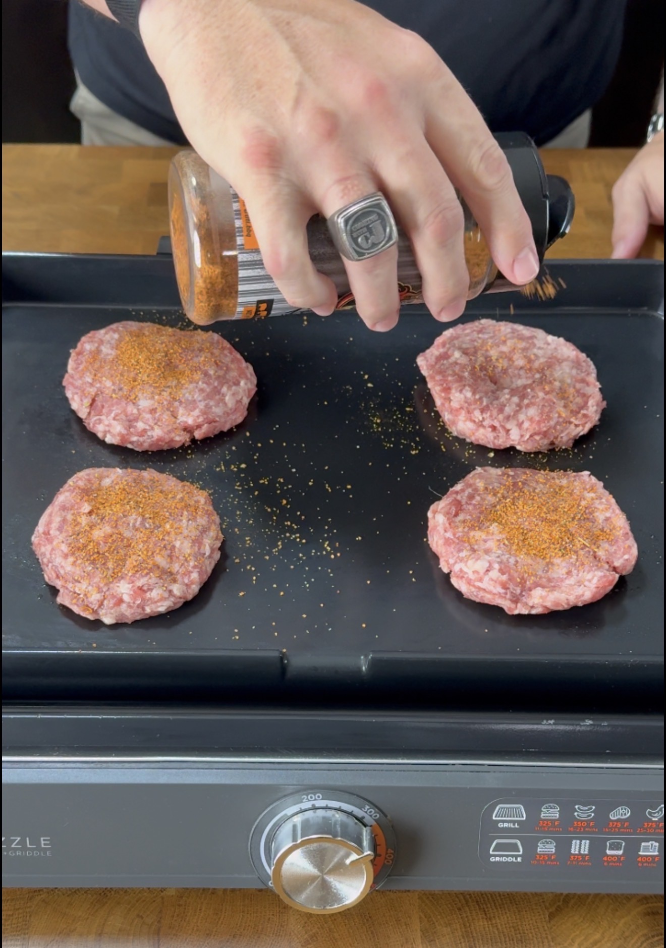 A hand sprinkles seasoning over four raw ground meat patties cooking on a black griddle with a silver temperature knob in front.