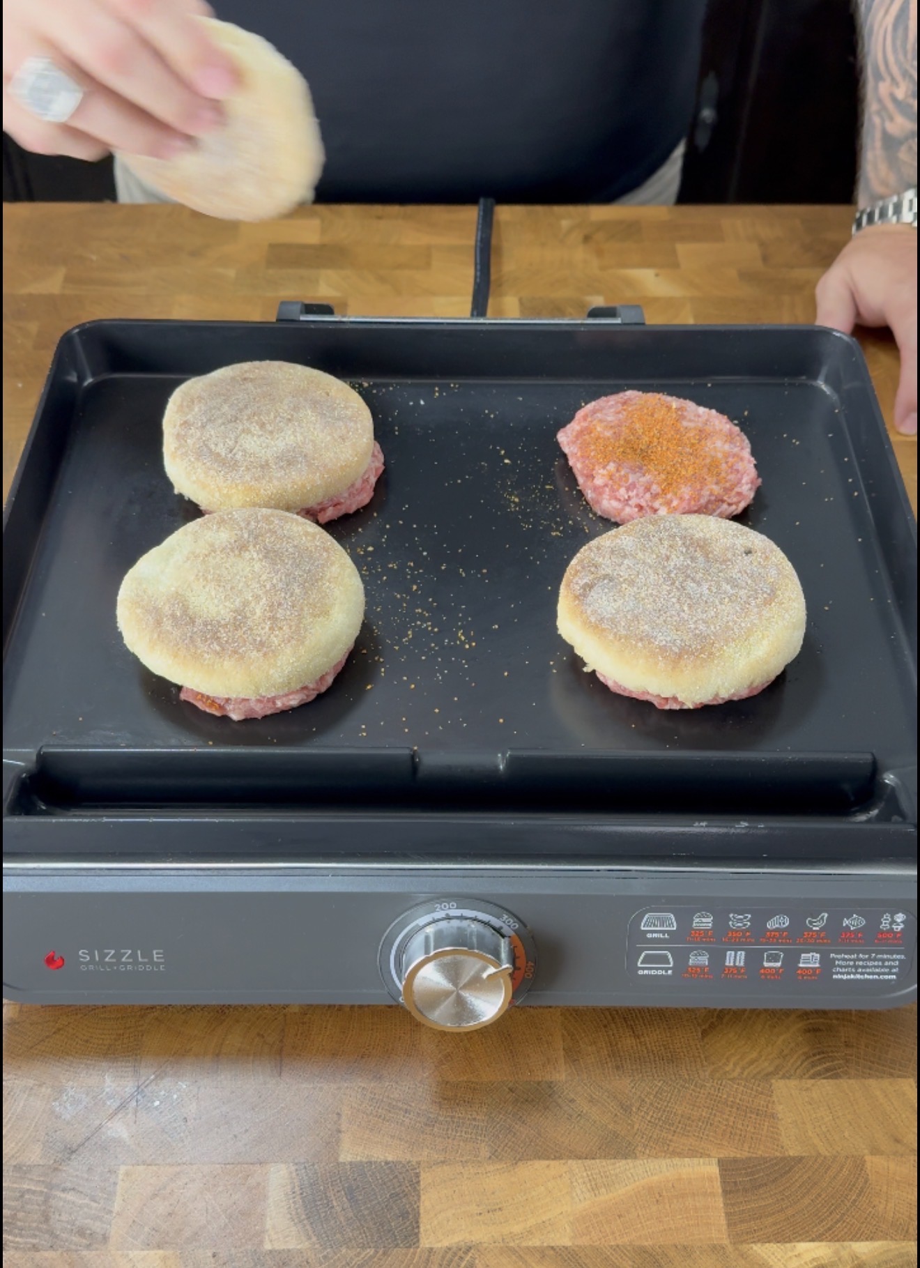 A person places an English muffin and raw burger patty combo on a griddle with three others. The griddle sits on a wooden counter.