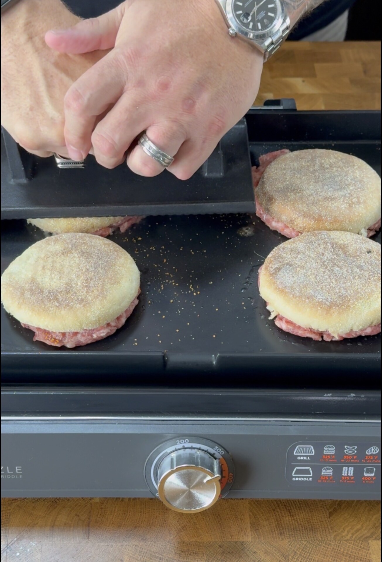 Two hands press a metal grill press onto English muffin breakfast sandwiches cooking on a griddle, with four sandwiches visible and a temperature dial in the foreground.