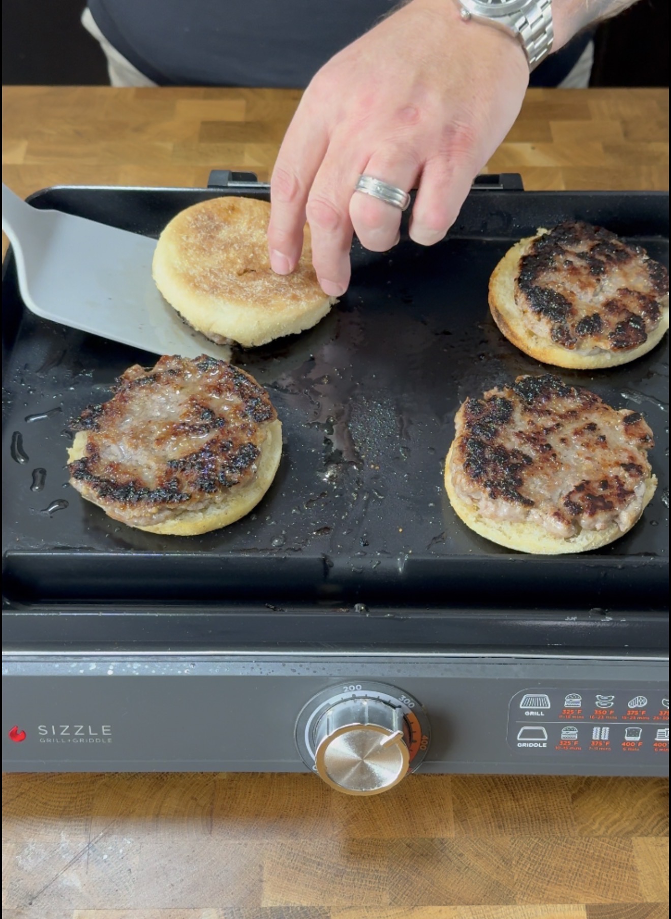 A hand with a ring uses a spatula to cook sausage patties on English muffins on a griddle, with a stainless steel control knob and cooking icons visible on the griddles front panel.