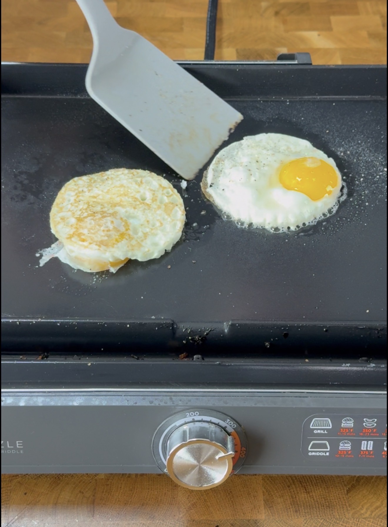 A spatula flips a fried egg on a black griddle next to another egg that is still cooking, with a temperature dial visible at the front of the griddle.