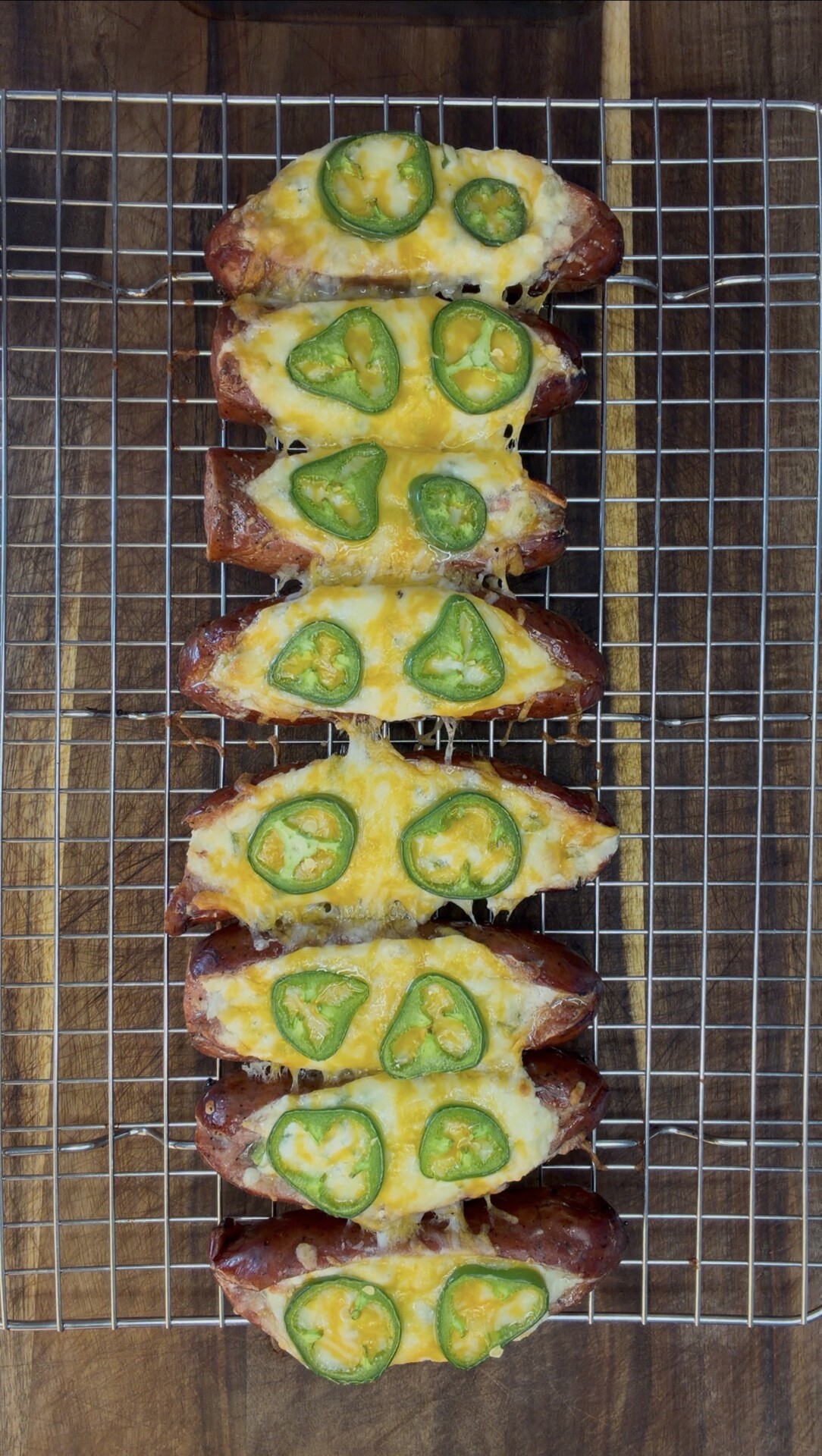 Six sausage halves topped with melted cheese and sliced jalapeños are arranged in two rows on a wire cooling rack, set on a wooden surface.