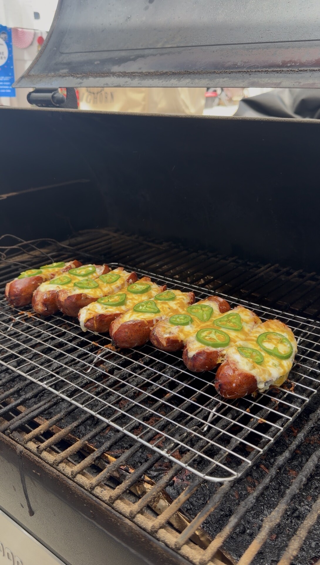 Several sausages topped with melted cheese and sliced jalapeños are being grilled on a wire rack inside a barbecue smoker.