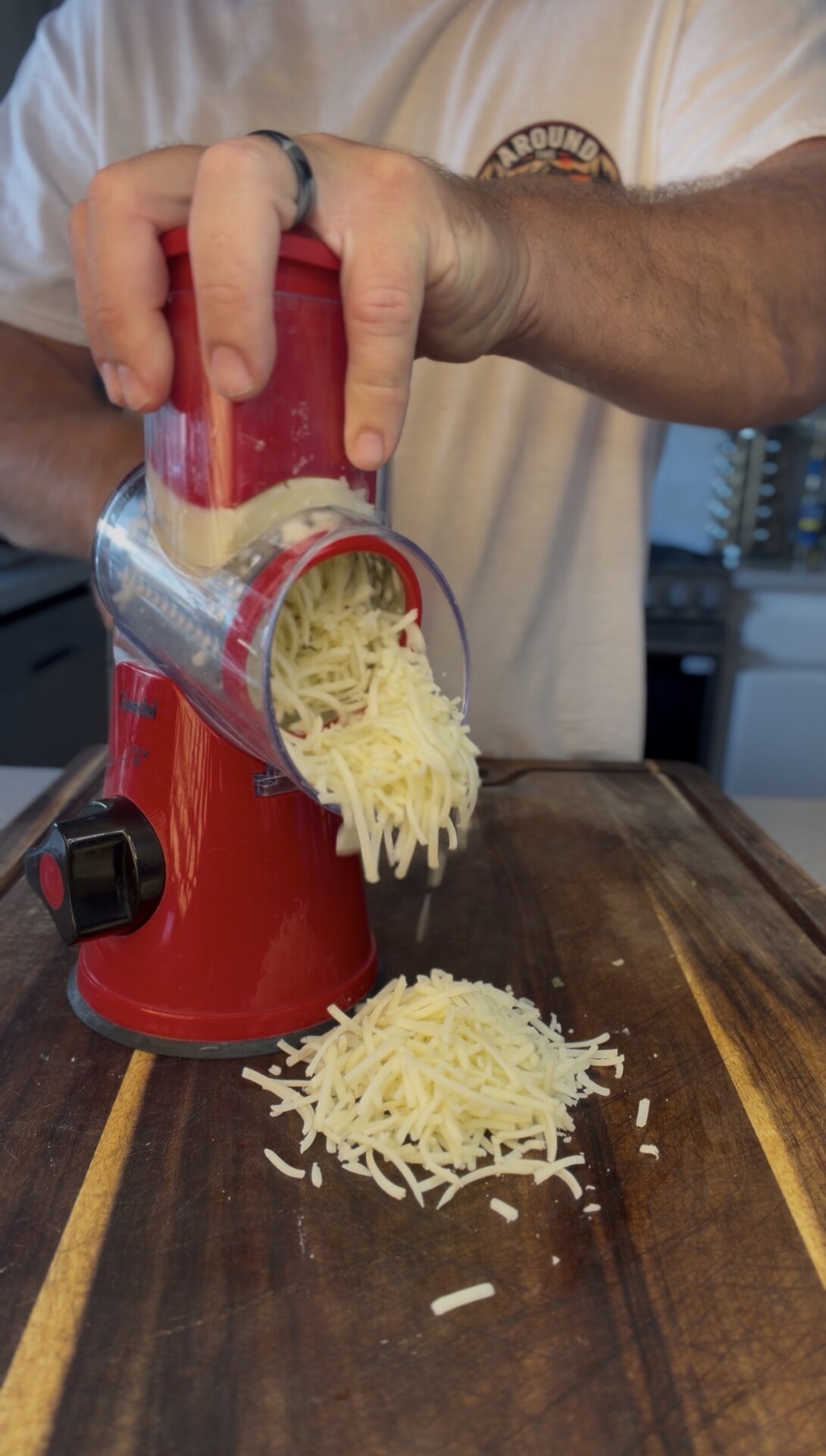 A person uses a red rotary cheese grater to shred cheese onto a wooden cutting board in a kitchen.