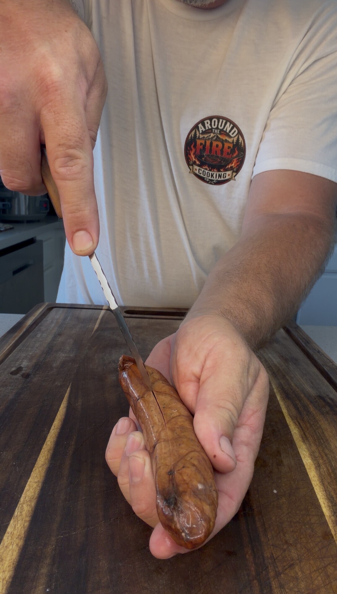 A person in a white Around the Fire T-shirt holds a sausage and slices it with a knife over a wooden cutting board.