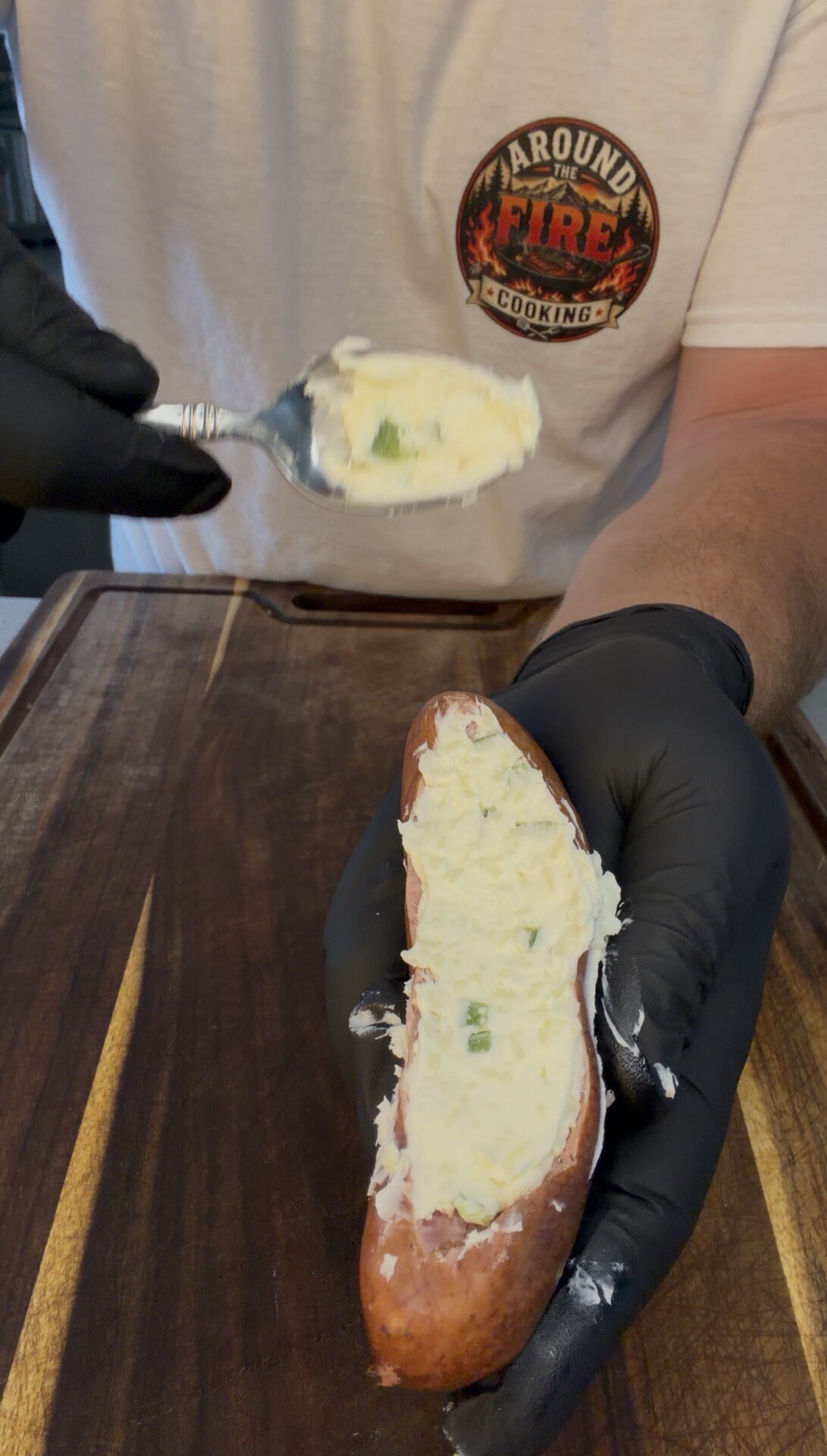 A person in black gloves fills a baked potato with creamy mashed potatoes and herbs, using a spoon. The person wears a white shirt with an Around the Fire Cooking logo.