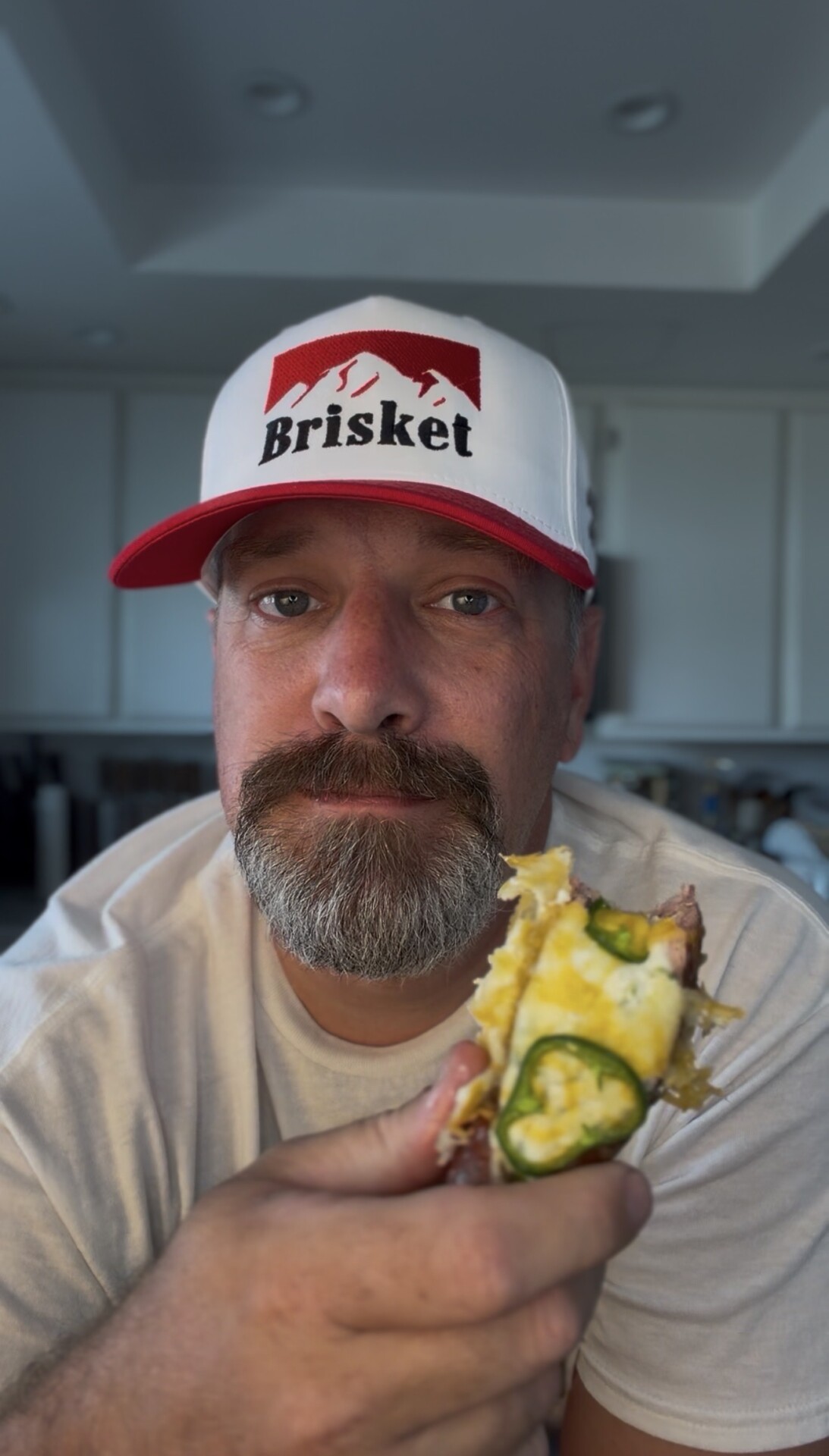 A man with a beard and mustache, wearing a white and red Brisket hat, holds up a cheesy dish topped with sliced jalapeños in a kitchen, looking directly at the camera.