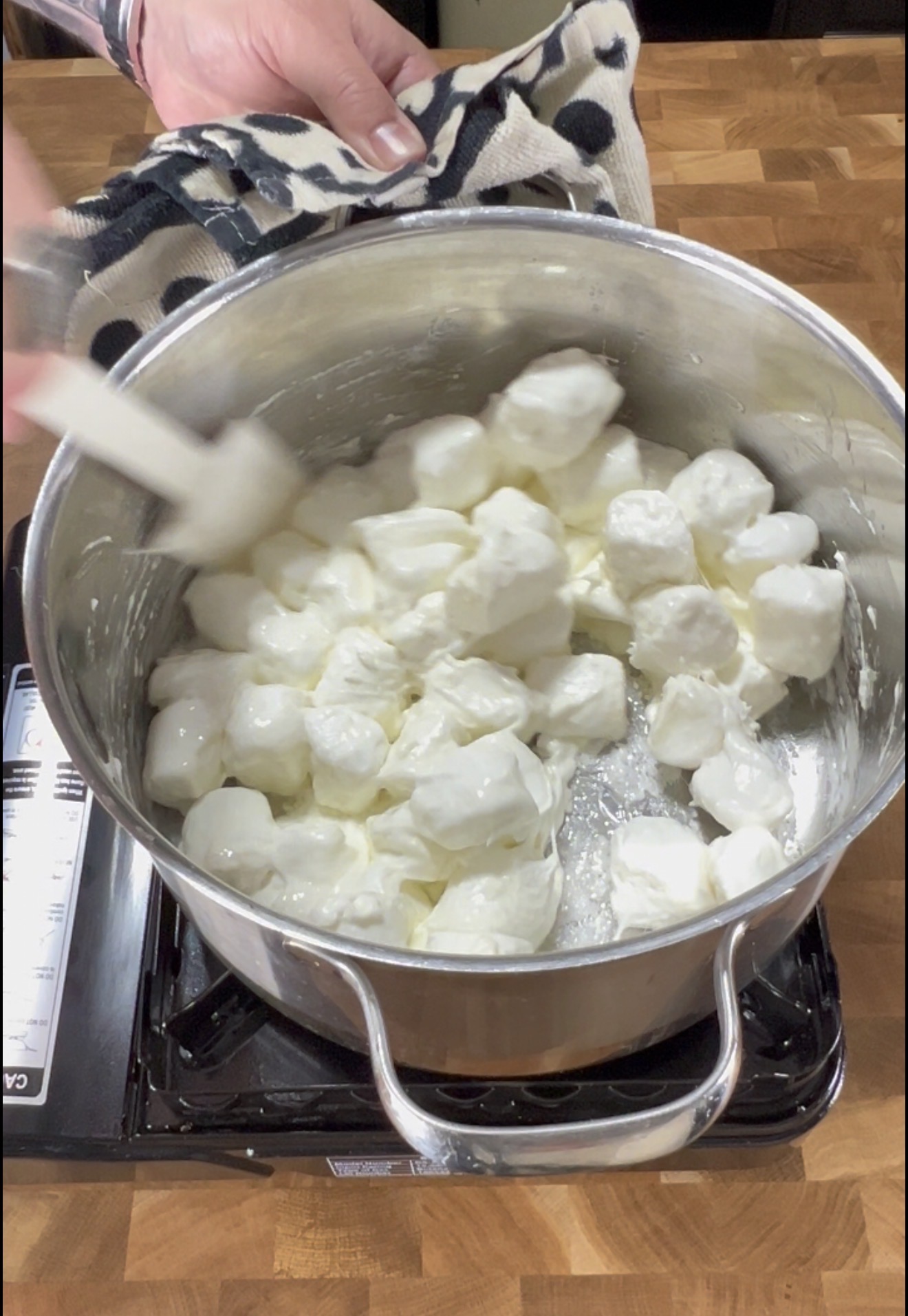 A hand stirs large white marshmallows melting in a metal pot on a stove, with a black and white towel nearby on a wooden countertop.