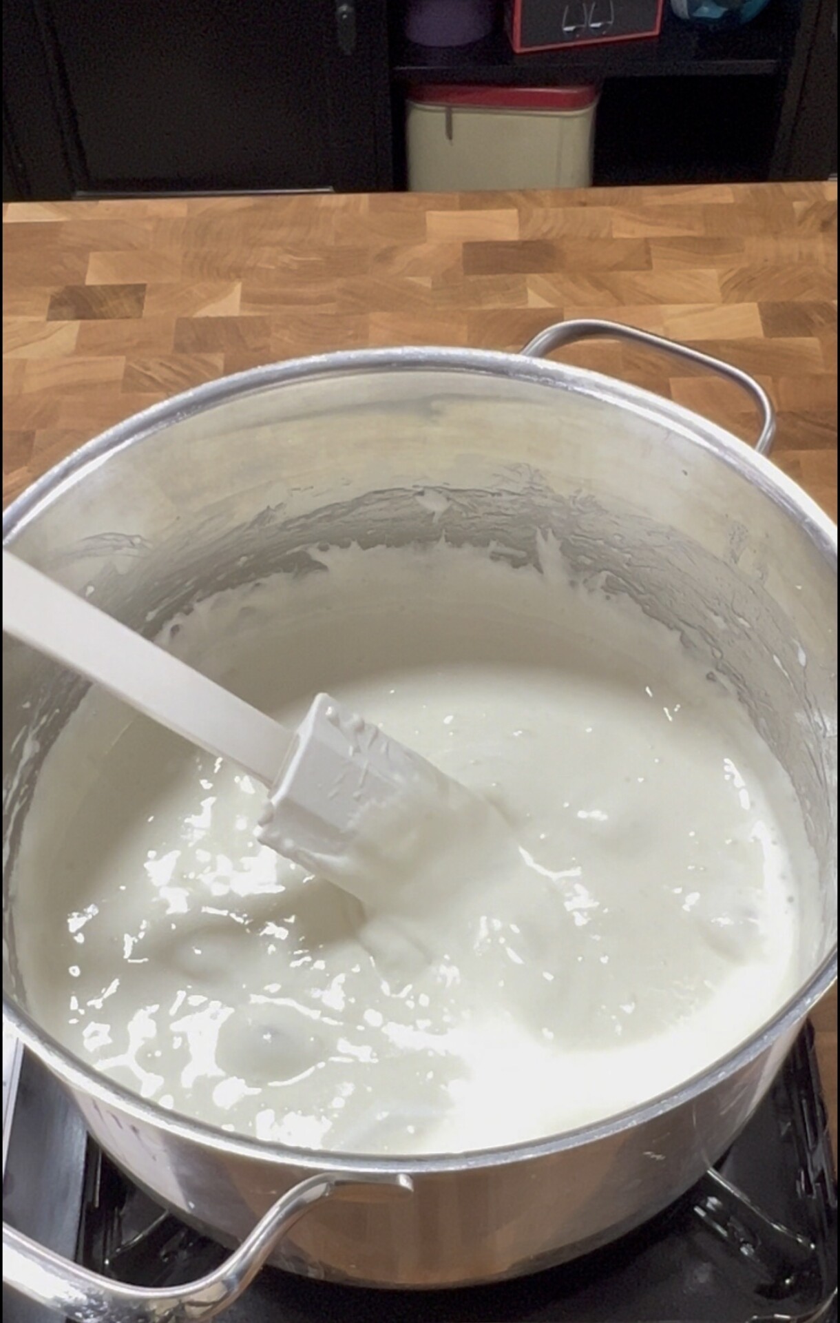 A metal pot filled with thick white liquid being stirred with a white spatula, sitting on a stovetop with a wooden countertop in the background.