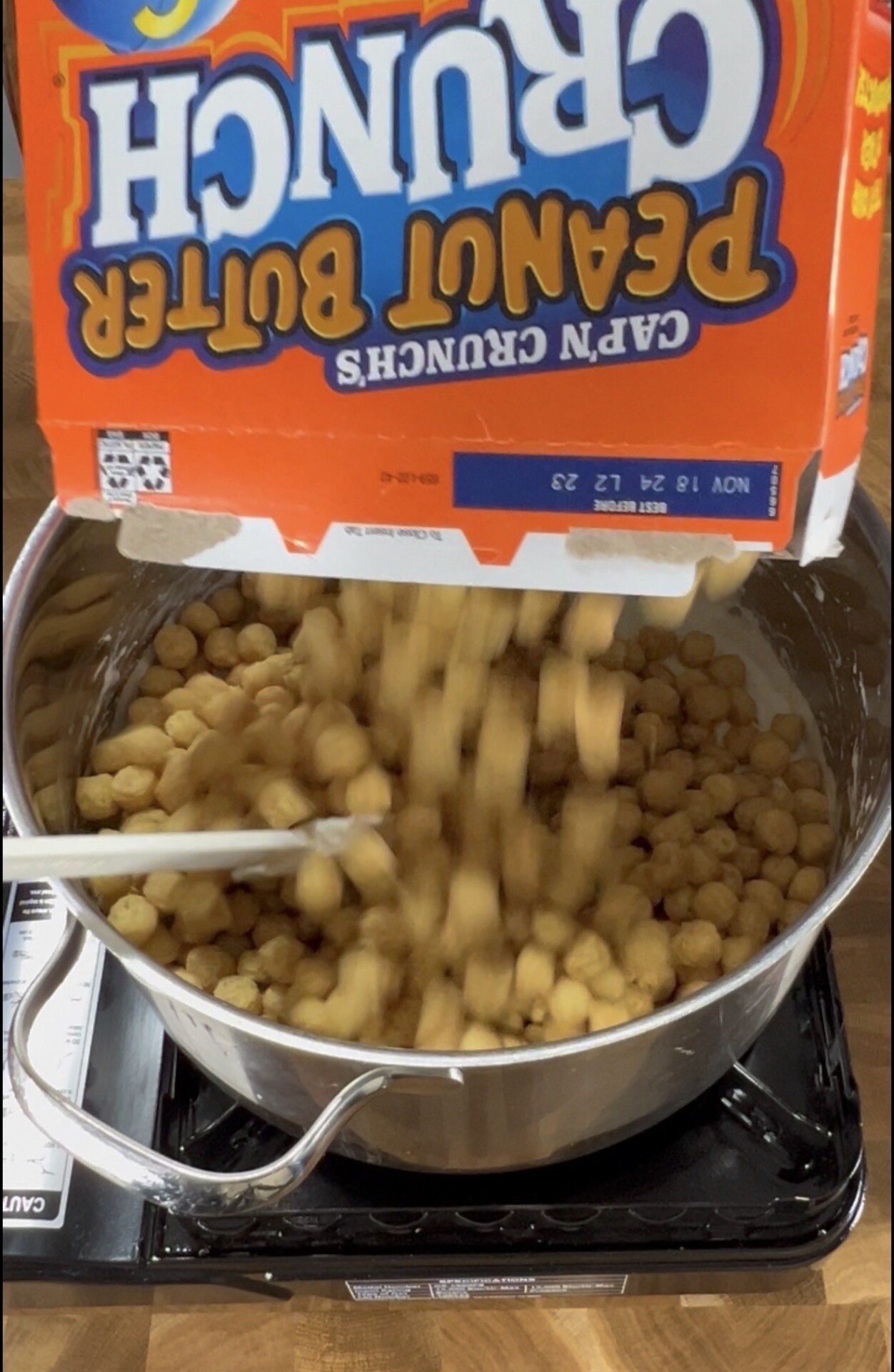 A box of Capn Crunchs Peanut Butter Crunch cereal is being poured into a large pot on a stove, with a spoon inside the pot.