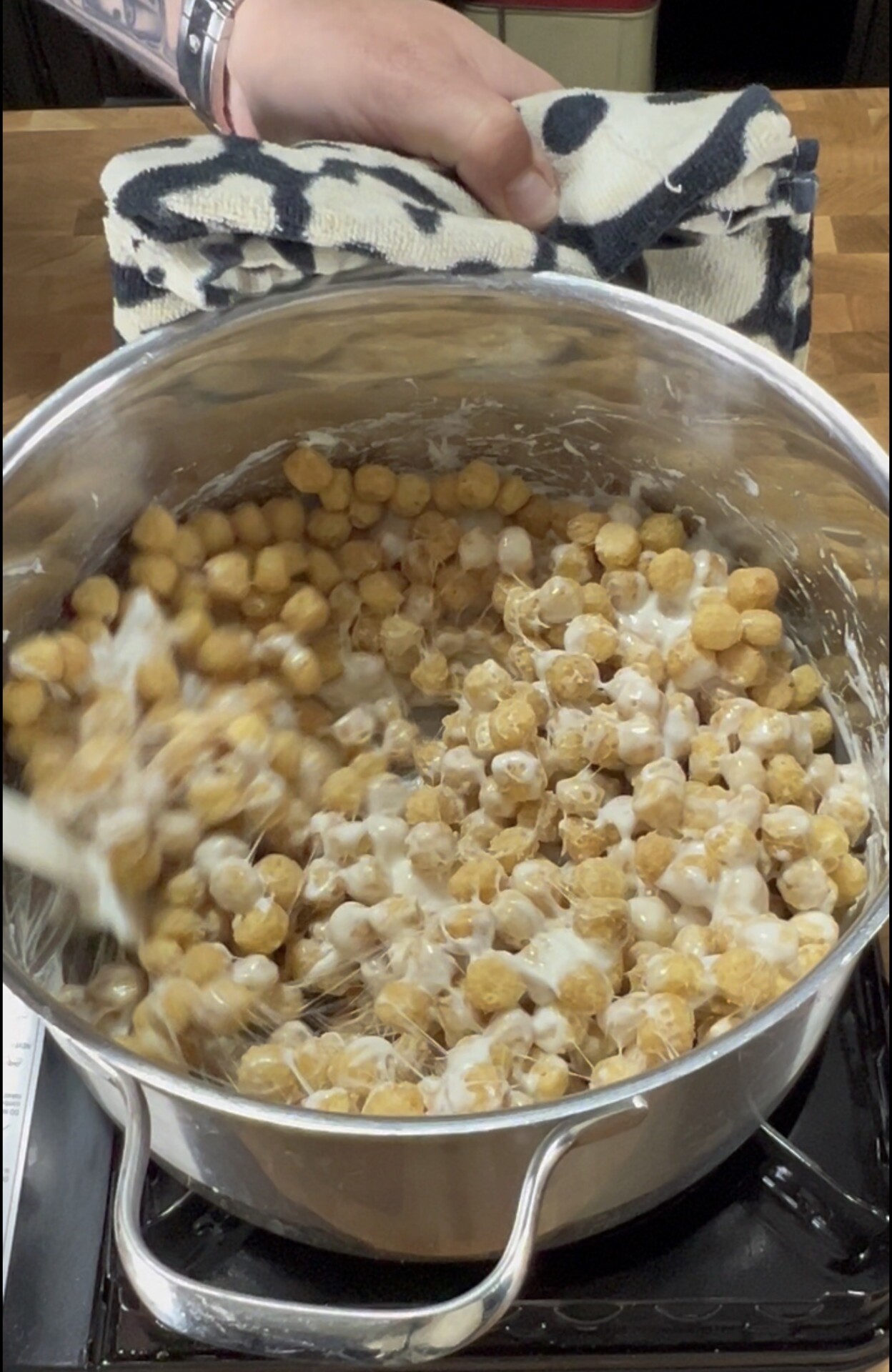 A hand holding a towel grips a pot handle as a spoon stirs cereal and melted marshmallows in a metal pot on a stovetop.