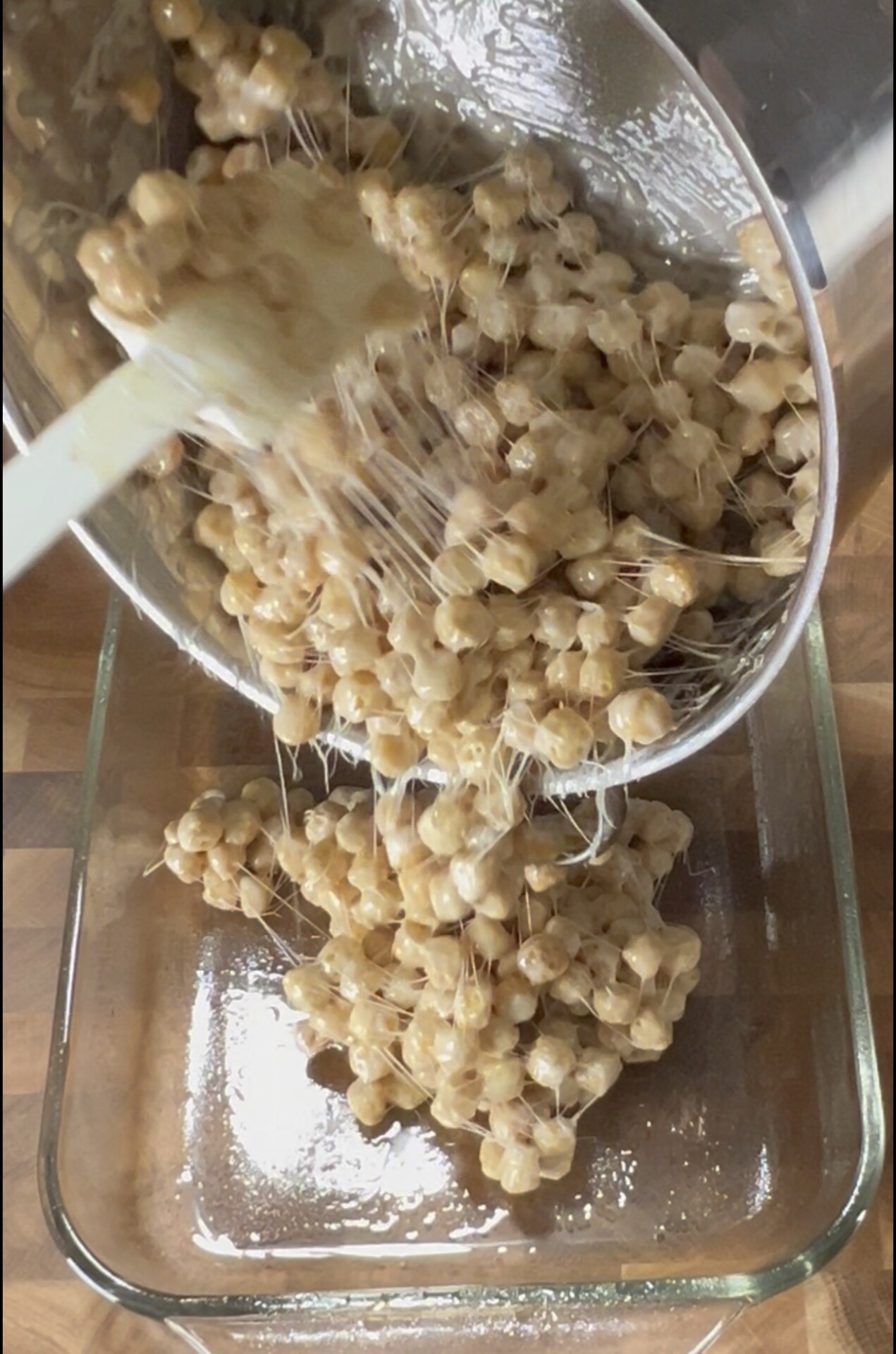 A spatula scrapes a sticky mixture of marshmallows and cereal from a pot into a glass baking dish on a wooden surface.