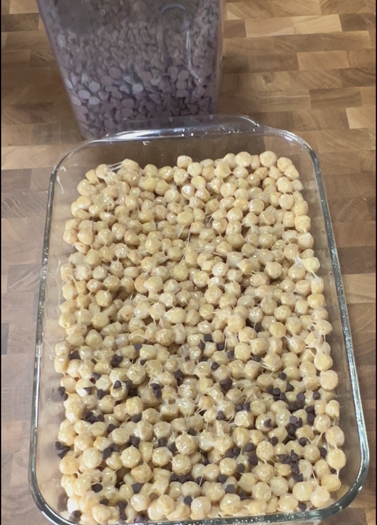 A glass baking dish filled with a cereal and marshmallow mixture sits on a wooden surface, with a container of chocolate chips in the background.