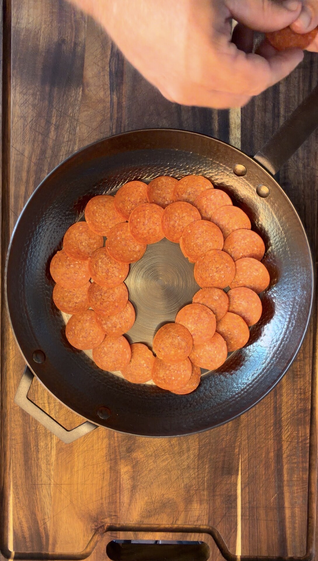 A hand arranges overlapping pepperoni slices in concentric circles inside a black skillet on a wooden surface.