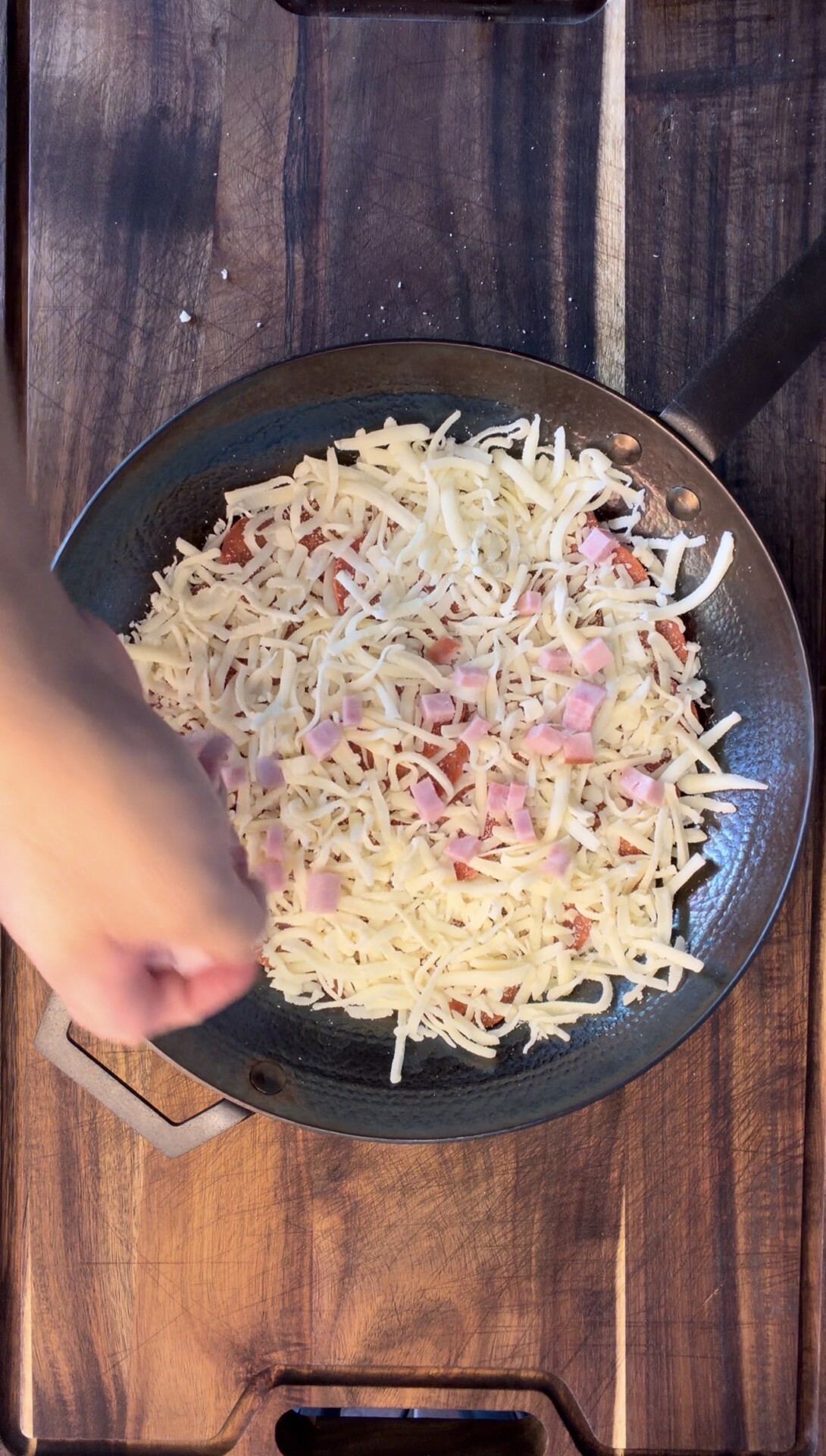A hand sprinkles diced ham onto a pizza in a black skillet topped with shredded cheese, on a wooden surface.