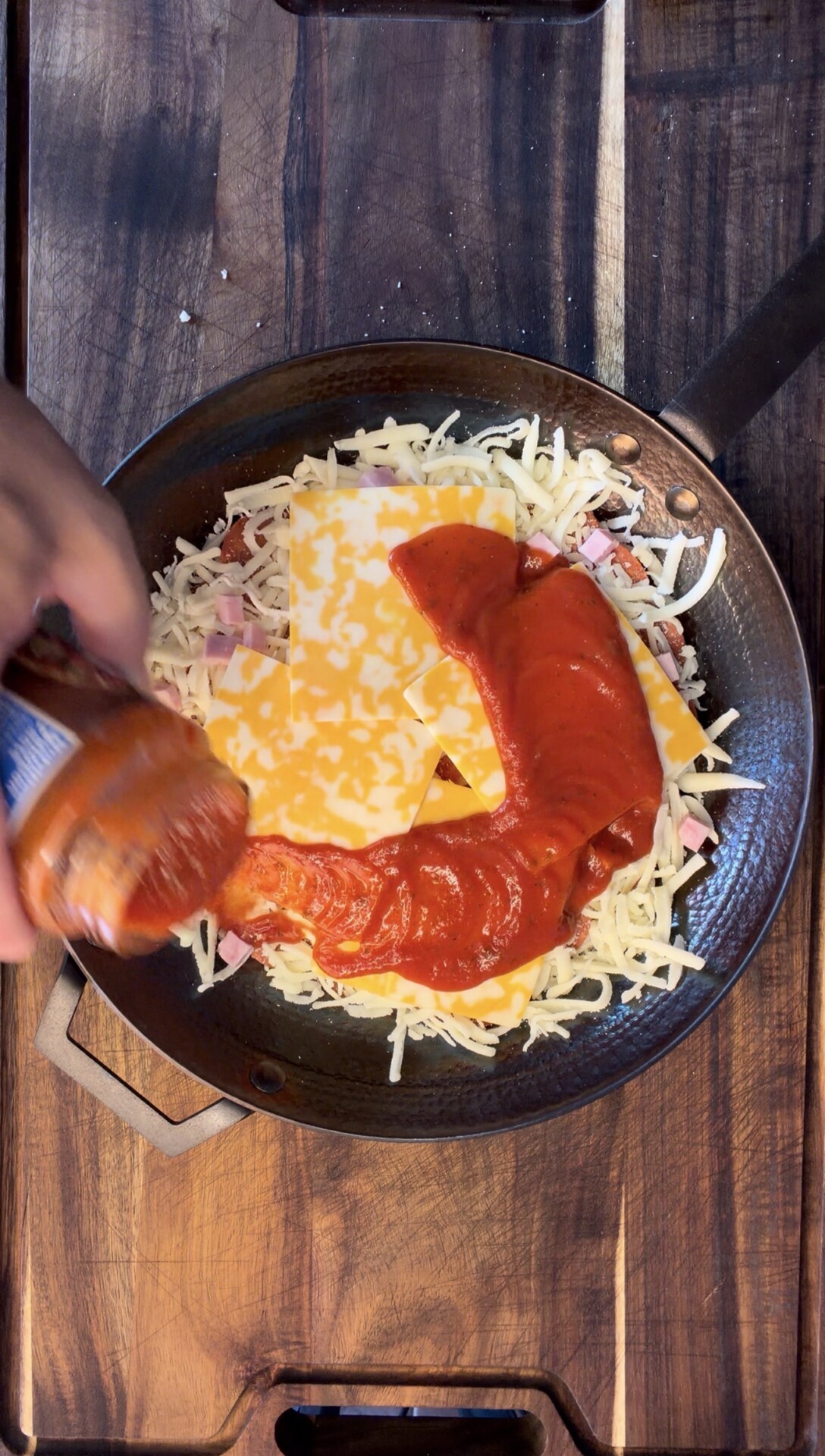 A hand pours pizza sauce from a jar onto cheese, ham, and shredded mozzarella in a skillet on a wooden surface.