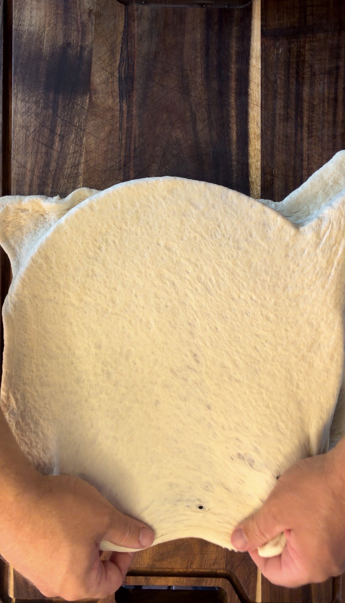 A person stretches pizza dough by hand on a dark wooden surface, preparing it for toppings.