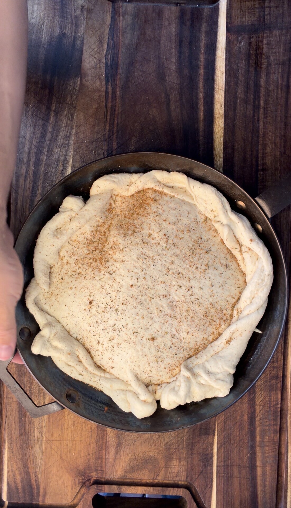A hand holds a cast iron skillet with an unbaked, twisted-edge pie crust sprinkled with spices, resting on a dark wooden surface.