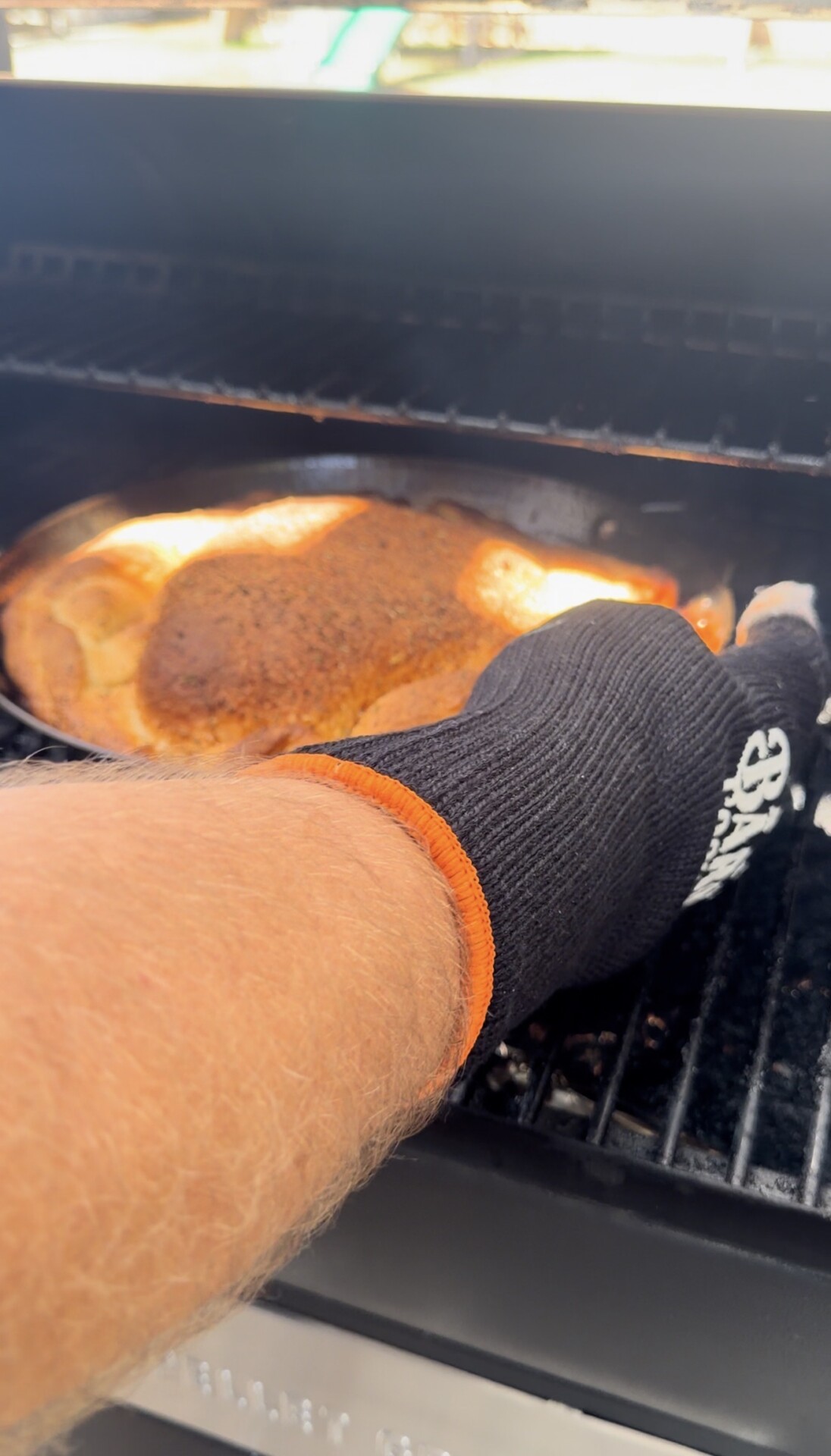 A person wearing a black grill glove is checking or adjusting a large, seasoned piece of meat cooking on a grill or smoker.