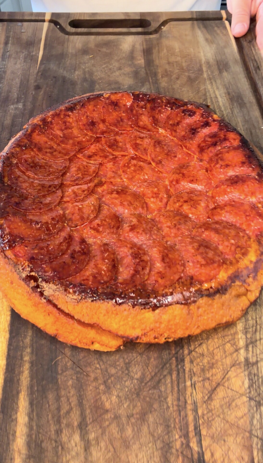 A round, golden-brown cake with a shiny, caramelized top sits on a wooden cutting board. One hand is visible holding the board.