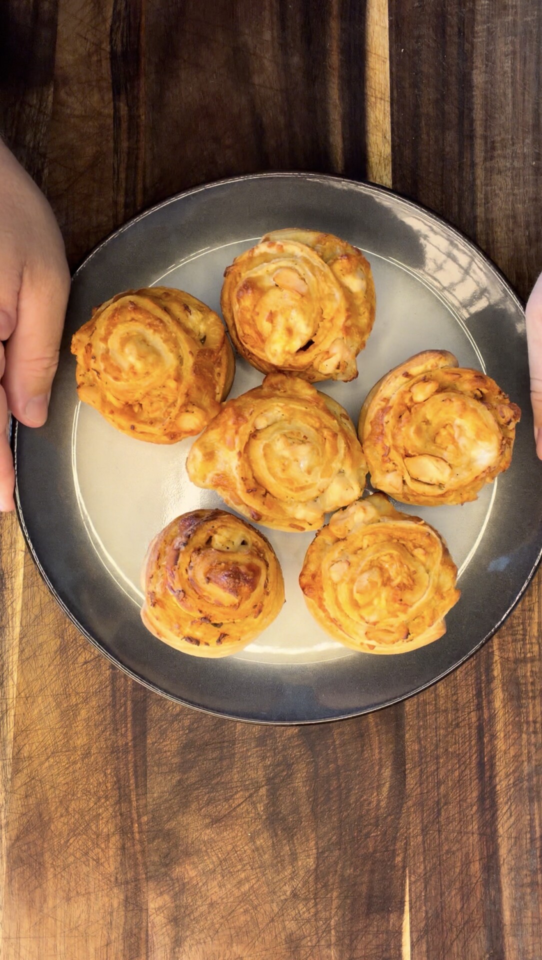 Five golden brown, spiral-shaped pastries are arranged on a white plate with a dark rim. The plate is held by two hands and placed on a wooden surface.