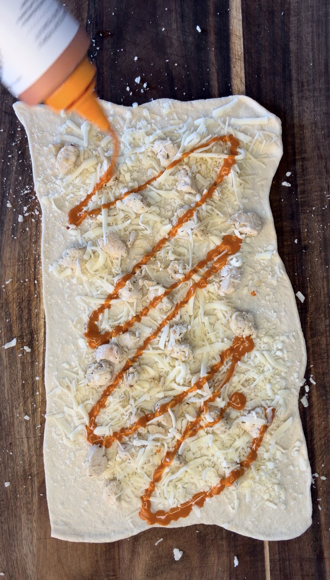 A rectangular piece of dough topped with shredded cheese and bits of chicken is drizzled with an orange sauce from a squeeze bottle on a wooden surface.