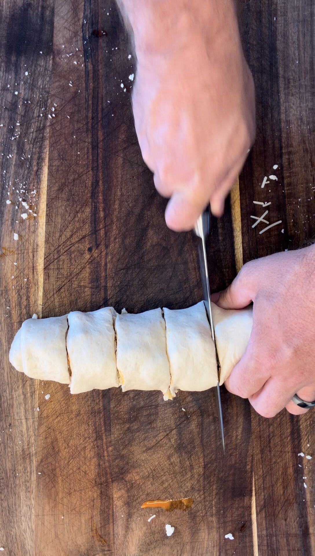 Hands slicing a rolled dough log into pieces on a wooden cutting board. The surface has some scattered flour and filling remnants.