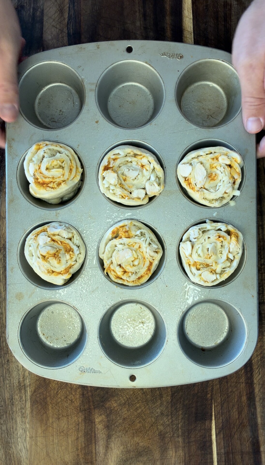 Hands holding a muffin tin with six unbaked pastry rolls, some topped with sliced almonds, in a wooden surface background. Three muffin cups are empty.