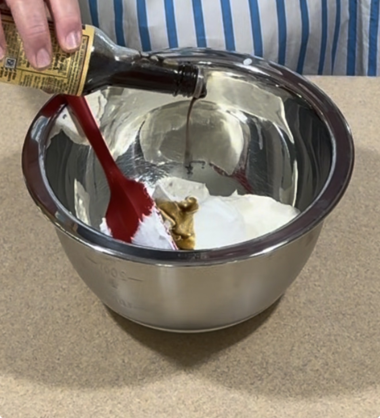 A hand pours vanilla extract into a metal bowl with white cream and brown ingredients, using a red spatula to mix. The person wears a blue and white striped apron.