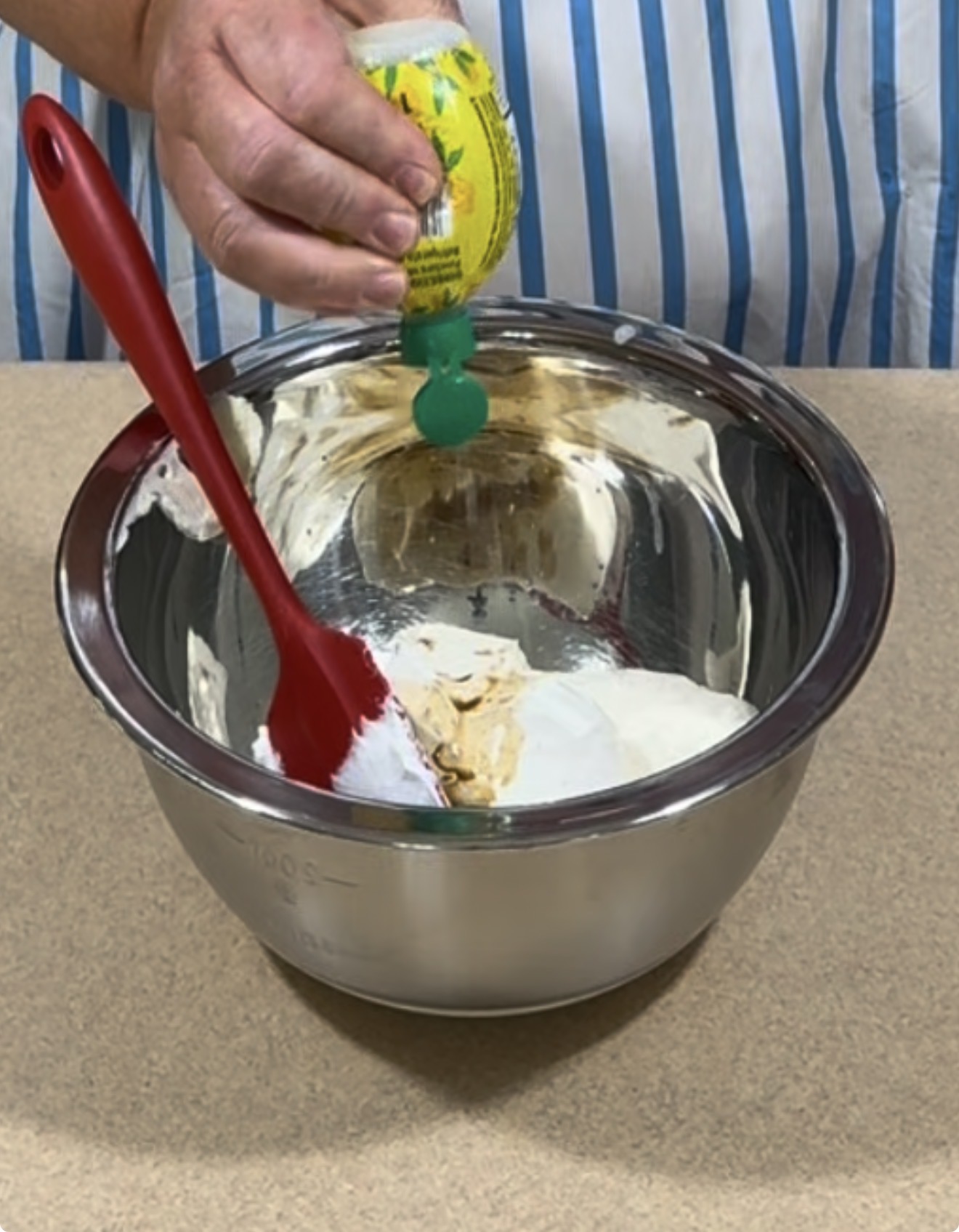 A person squeezes lemon juice from a yellow bottle into a metal mixing bowl with a red spatula and cream inside, on a countertop.