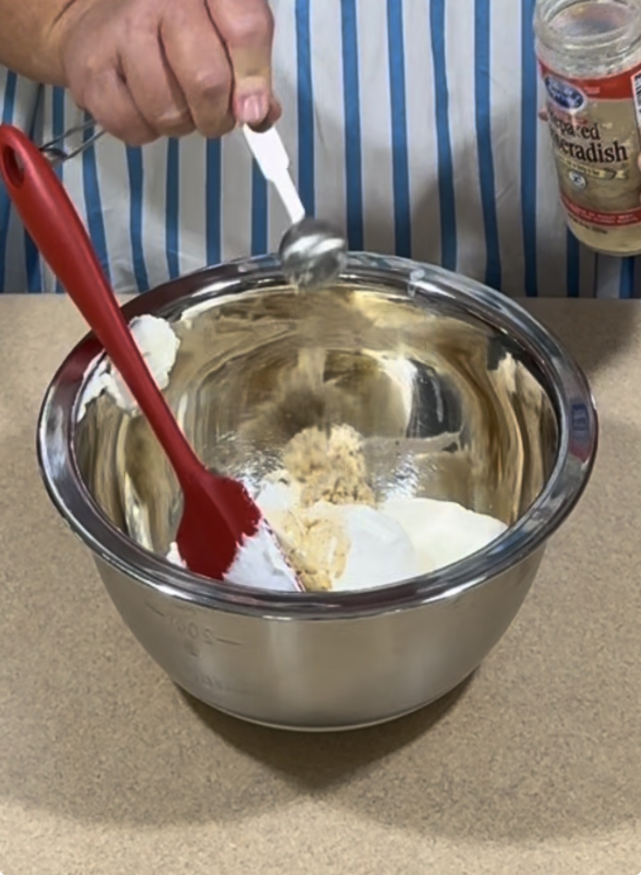 A person adds a spoonful of an ingredient to a metal mixing bowl with brown sugar and white mixture, holding a jar of horseradish nearby. A red spatula rests in the bowl on a countertop.