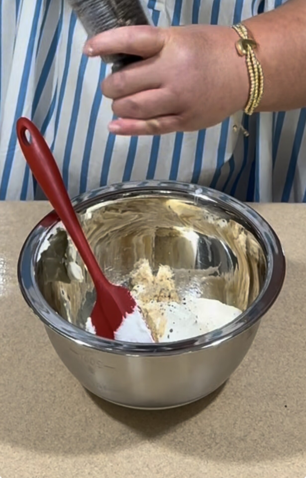 A person grinds pepper into a metal mixing bowl containing white and tan ingredients, with a red spatula resting inside. The person wears a striped shirt and gold bracelets.