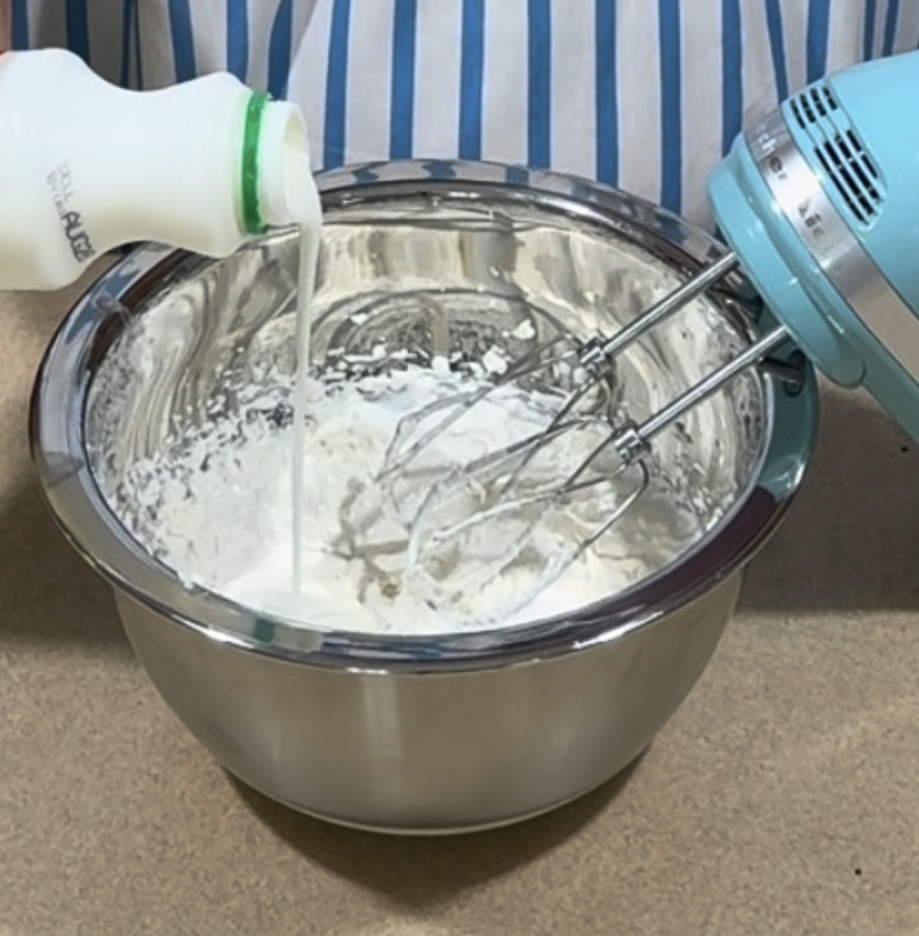 Person pouring milk into a mixing bowl with white powder while using a blue electric hand mixer; the person is wearing a blue and white striped apron.