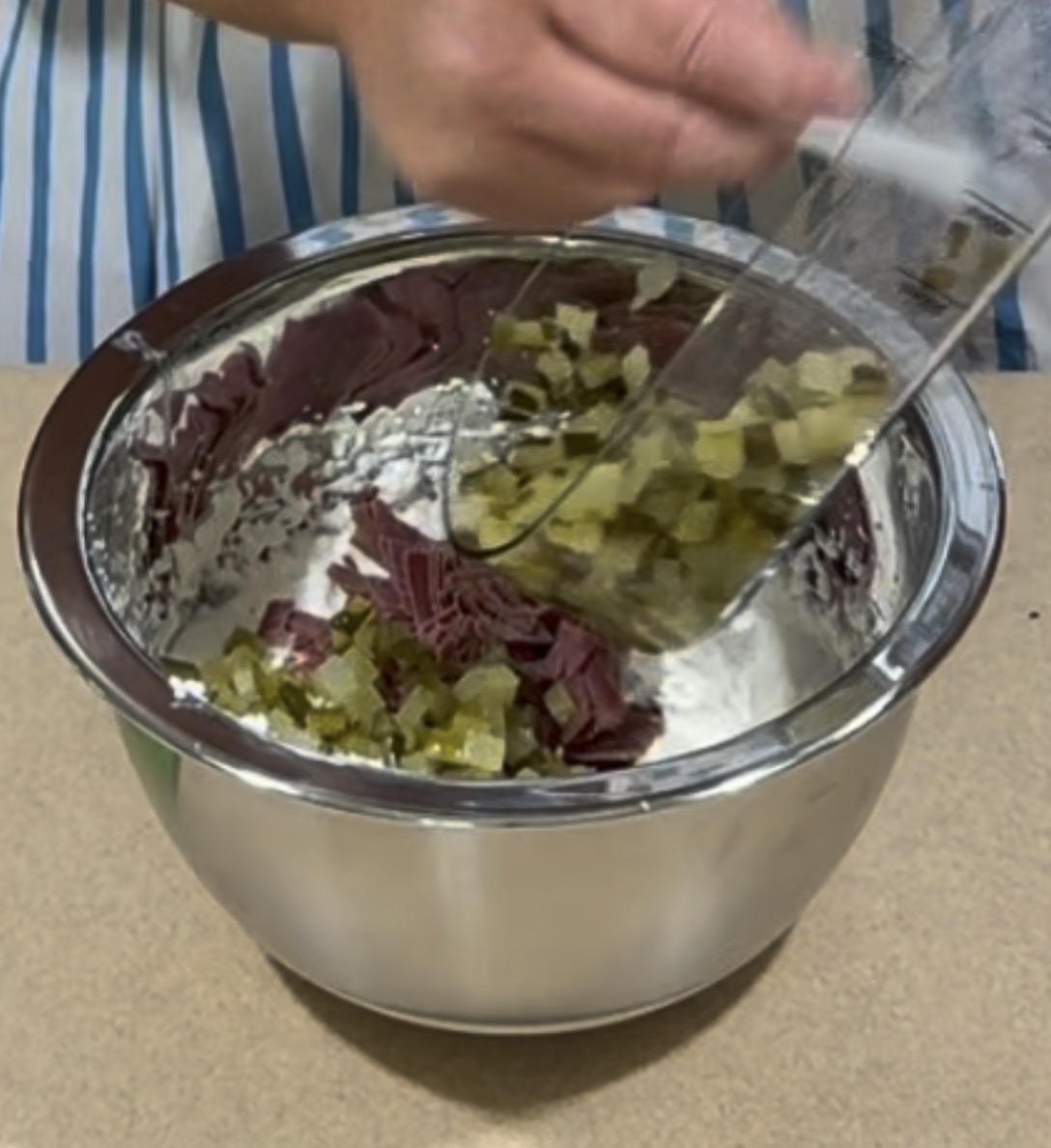 A person adds chopped pickles and meat into a mixing bowl containing a creamy mixture. The bowl is placed on a countertop, and the person is wearing a striped shirt.