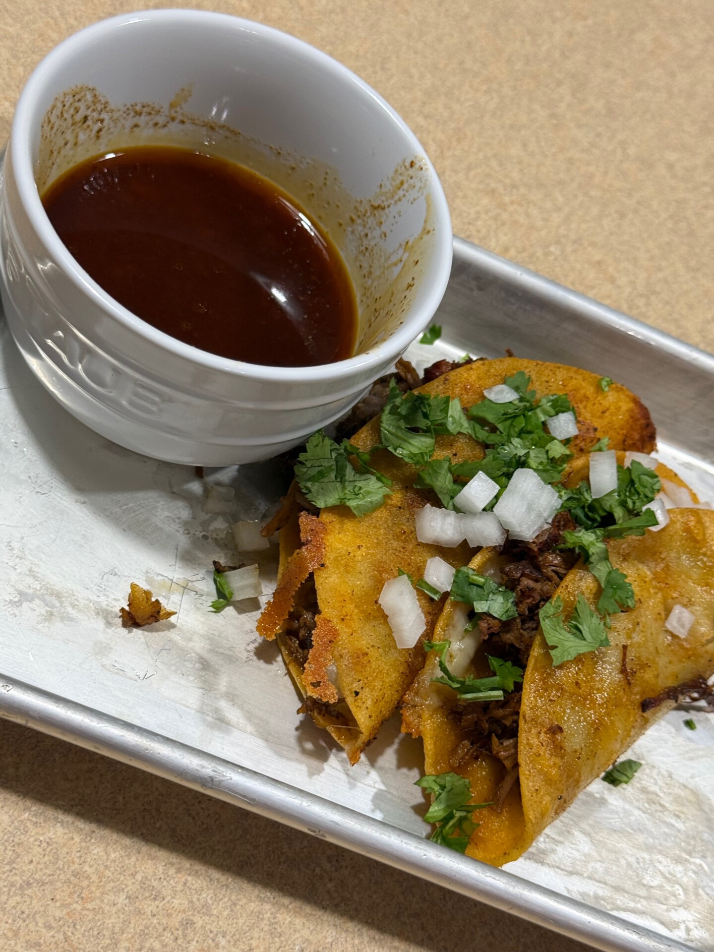Three birria tacos topped with chopped onions and cilantro sit on a metal tray next to a small white bowl filled with dark broth for dipping.