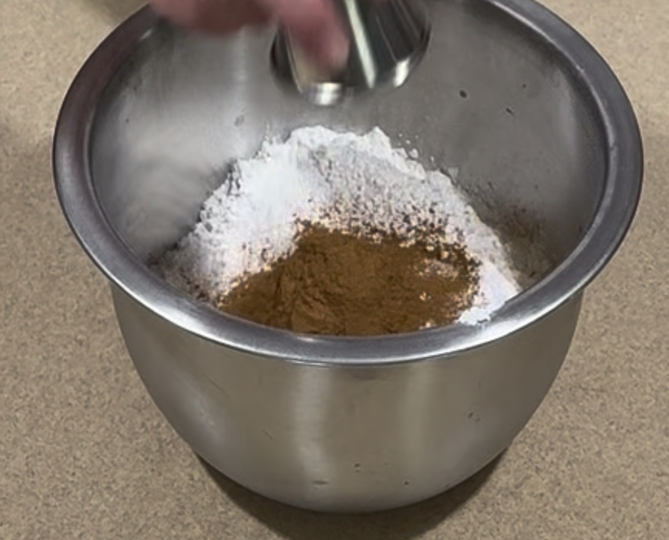 A hand pours an ingredient from a small metal cup into a mixing bowl containing flour and cinnamon, set on a brown countertop.