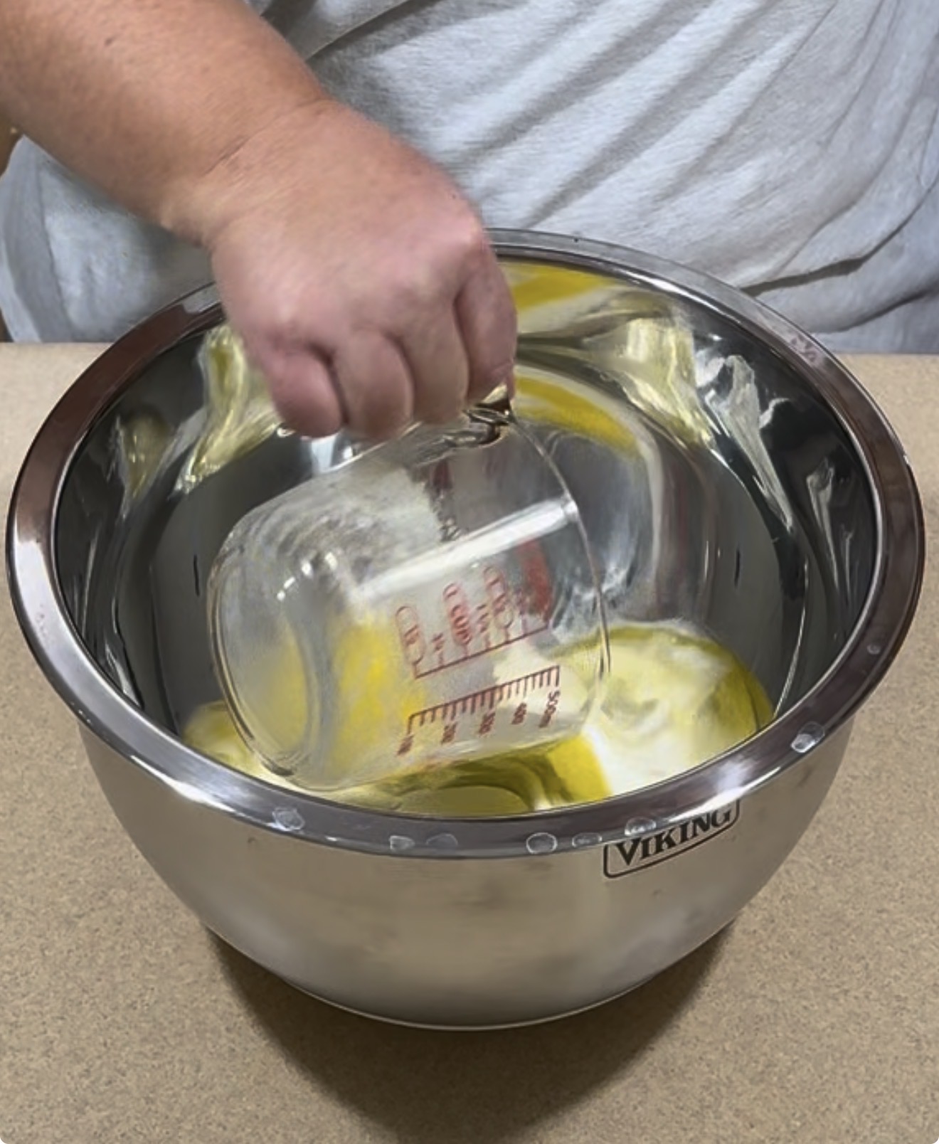 A person pours liquid from a glass measuring cup into a stainless steel mixing bowl containing a yellow mixture, set on a countertop.