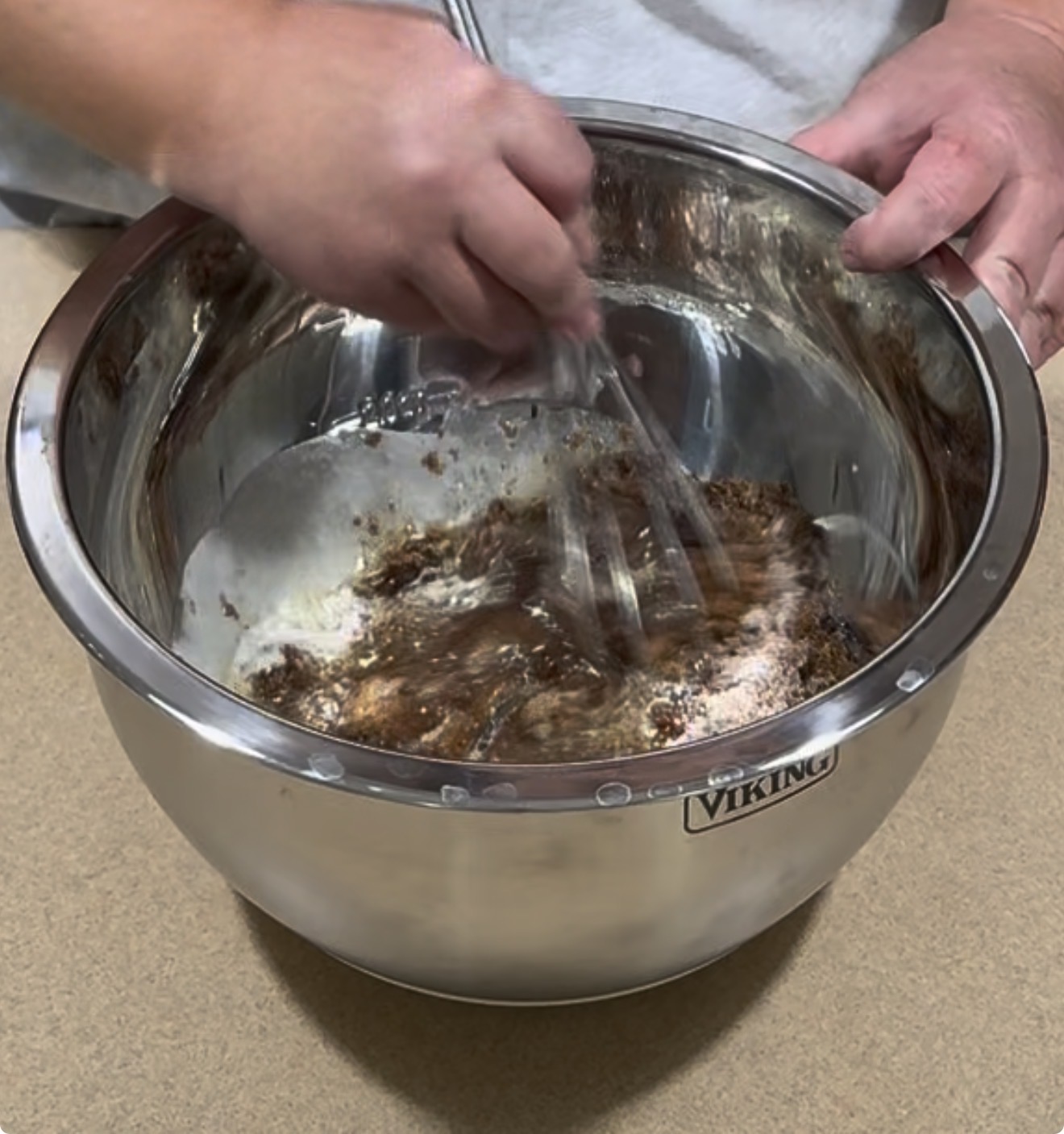 A person uses a whisk to mix chocolate batter in a large stainless steel bowl placed on a beige countertop.