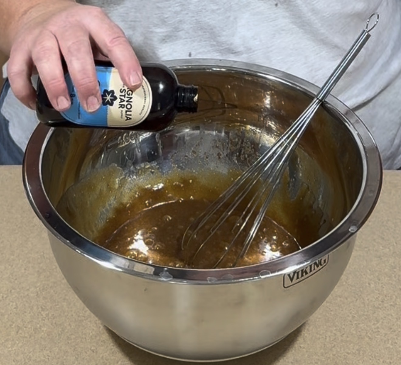 A person pours vanilla extract from a bottle into a stainless steel mixing bowl with brown batter, using a whisk to mix the ingredients.
